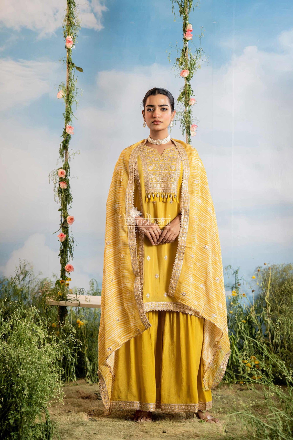Woman in a yellow traditional outfit standing on a swing with floral decorations against a blue sky.