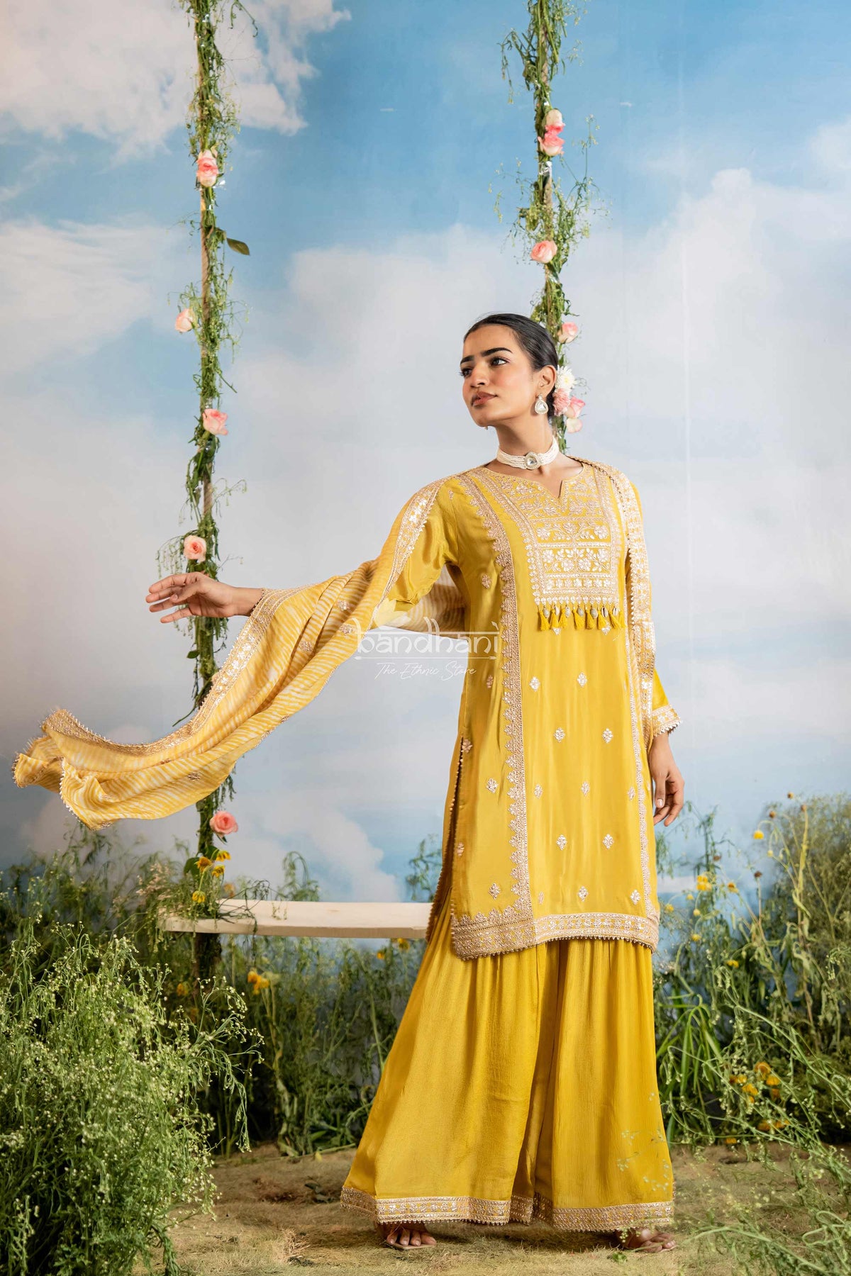 Woman in a yellow traditional outfit sitting on a swing with floral decorations against a blue sky.