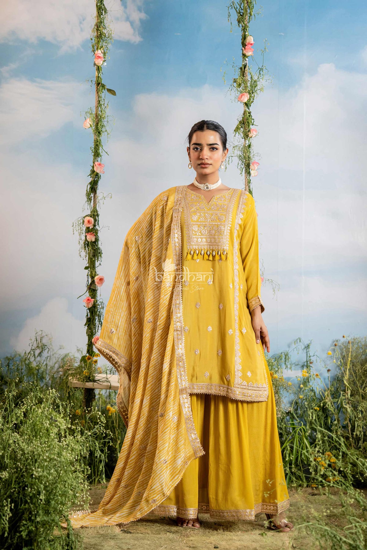 Woman in a yellow traditional outfit standing on a swing with floral decorations against a blue sky.