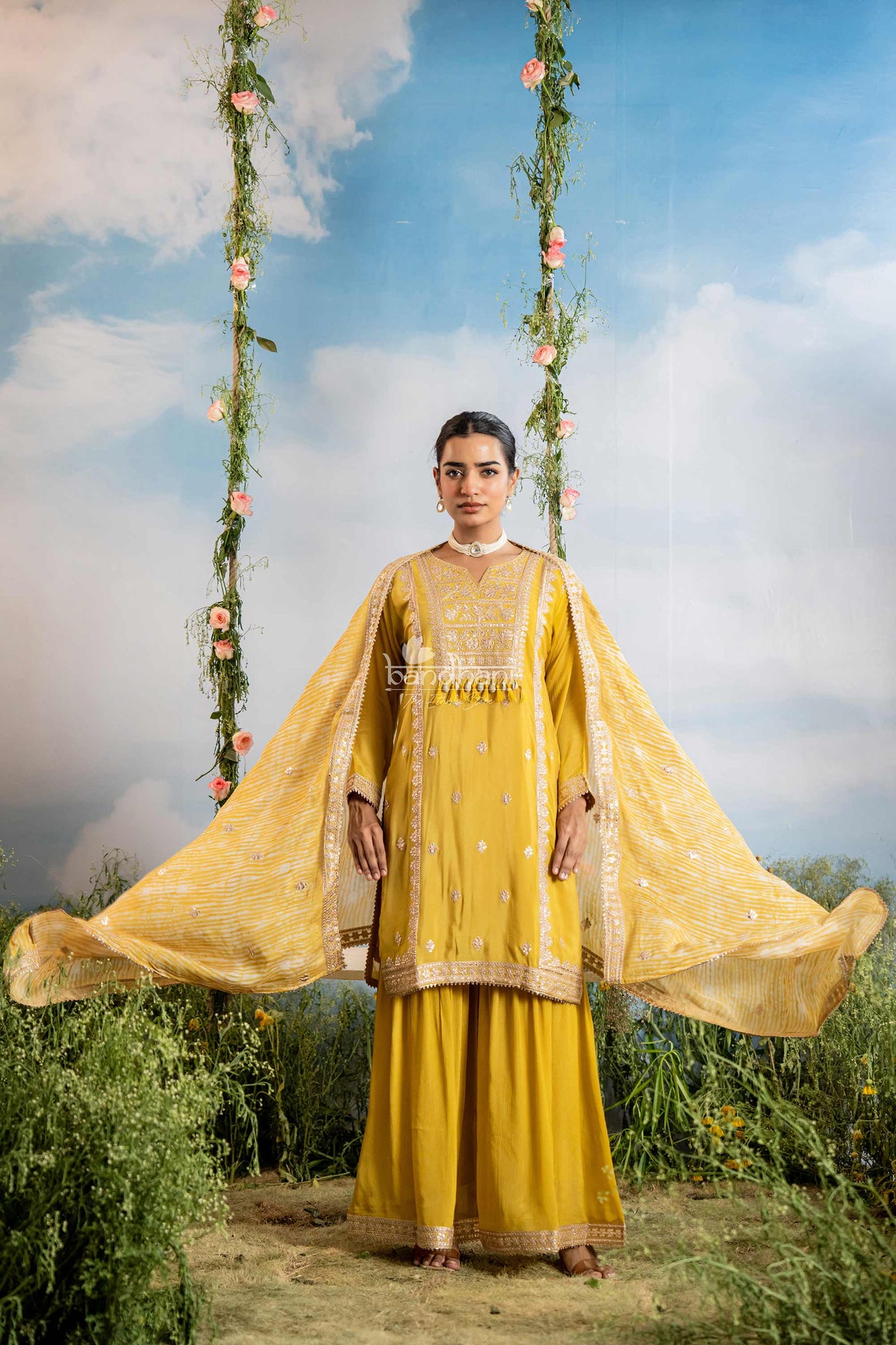Woman in a yellow traditional outfit standing on a swing with floral decorations against a blue sky.