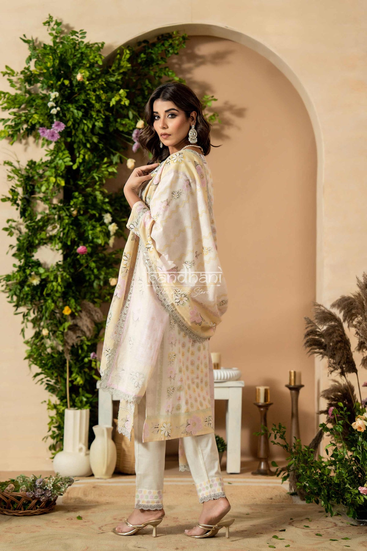 Woman in a traditional outfit standing in a decorated indoor setting with plants and candles.