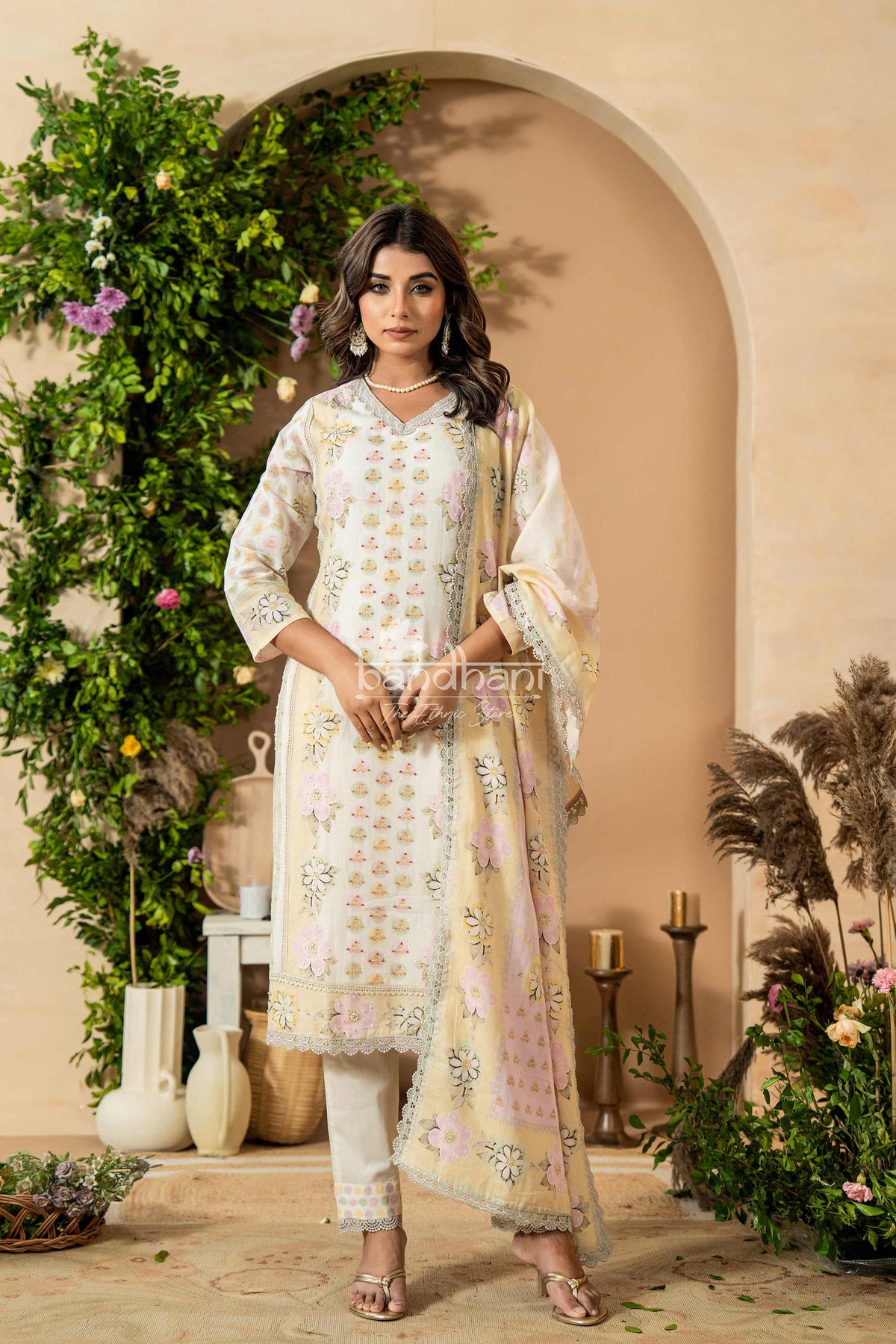 Woman in a traditional outfit standing in a decorative setting with plants and archway.