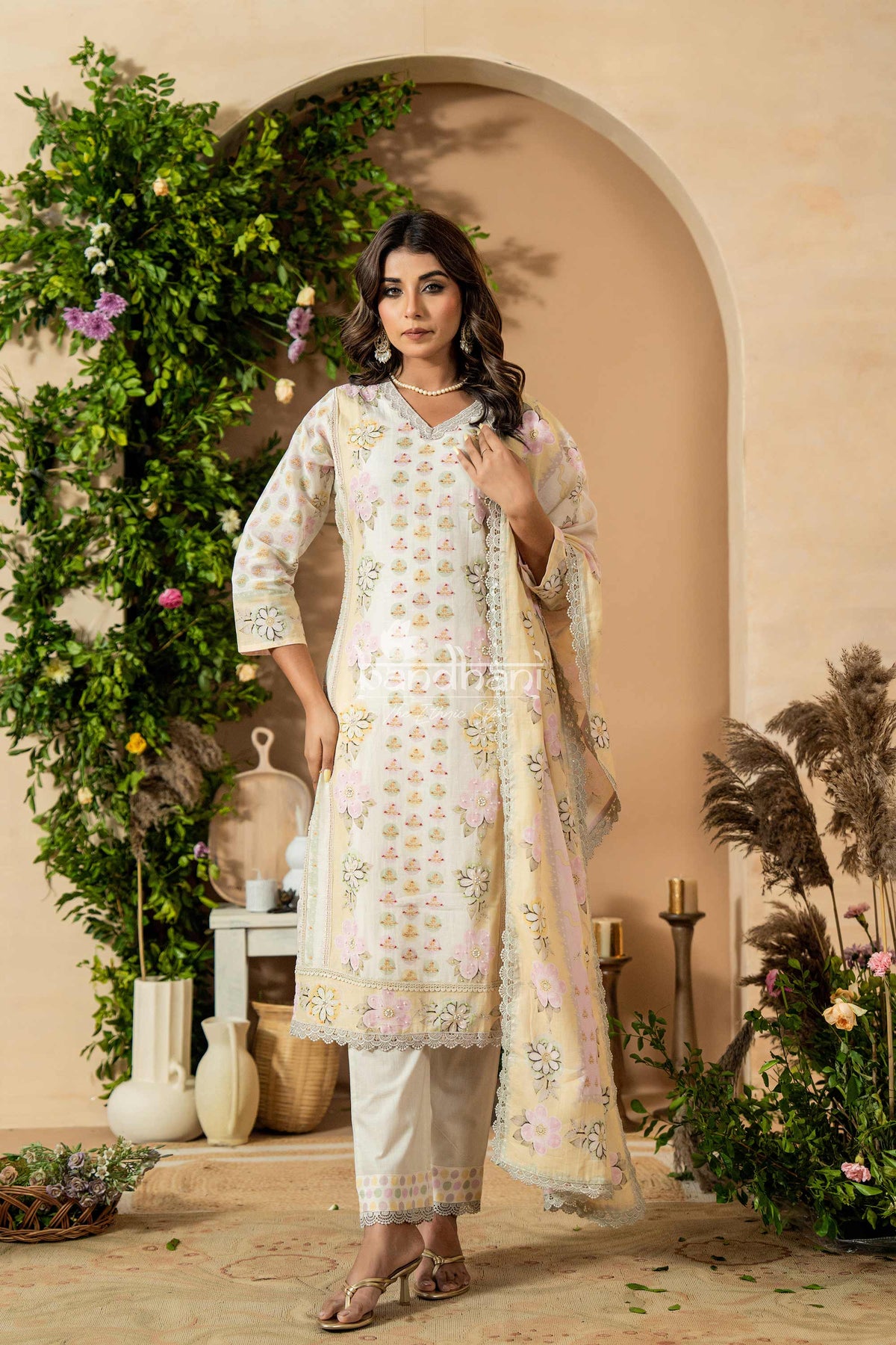 Woman in a traditional outfit standing in a decorative setting with plants and archway.