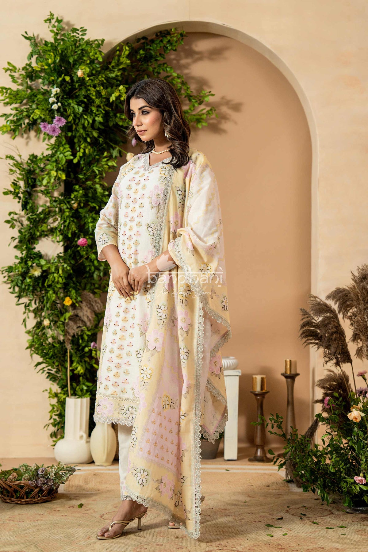 Woman in a traditional outfit standing in a decorated indoor setting with plants and candles.