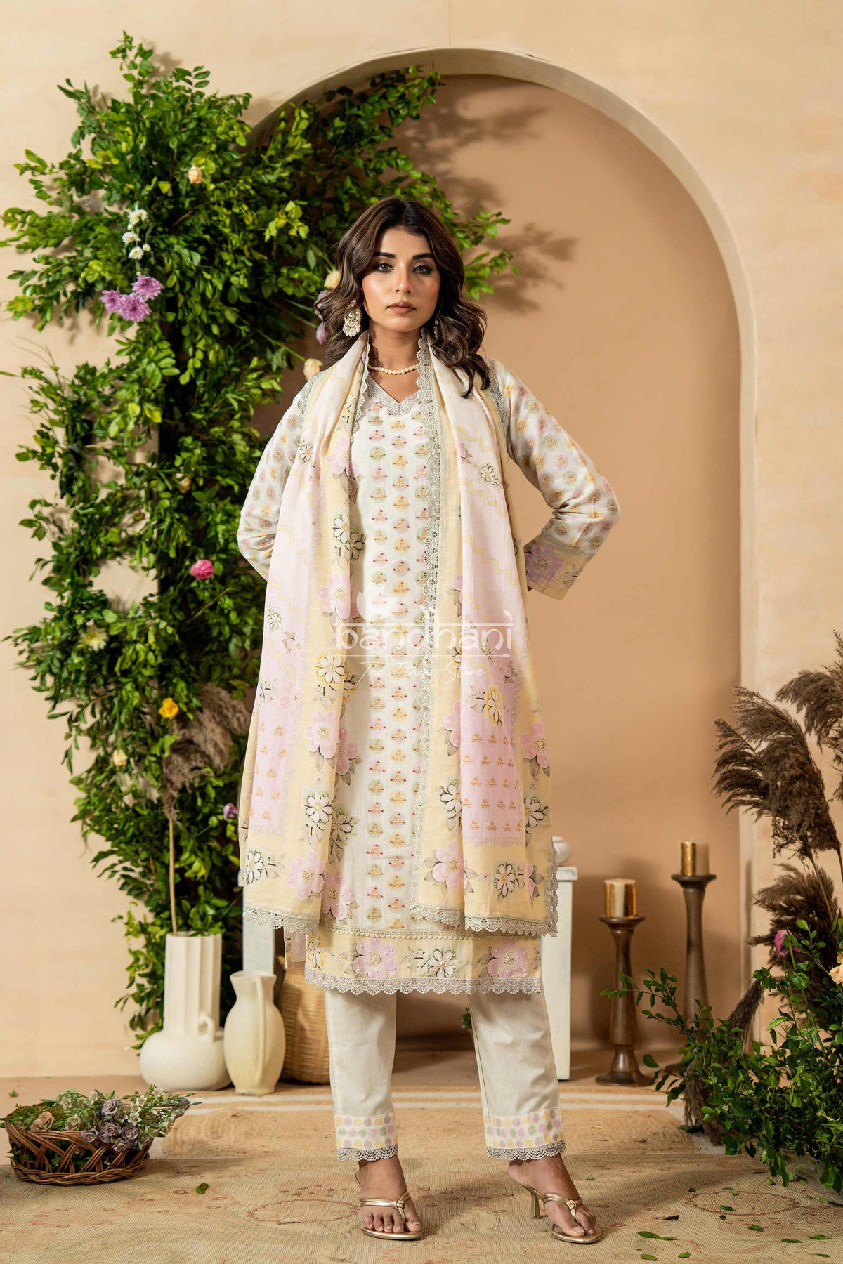 Woman in a traditional outfit with floral patterns standing in front of decorative plants and archway.
