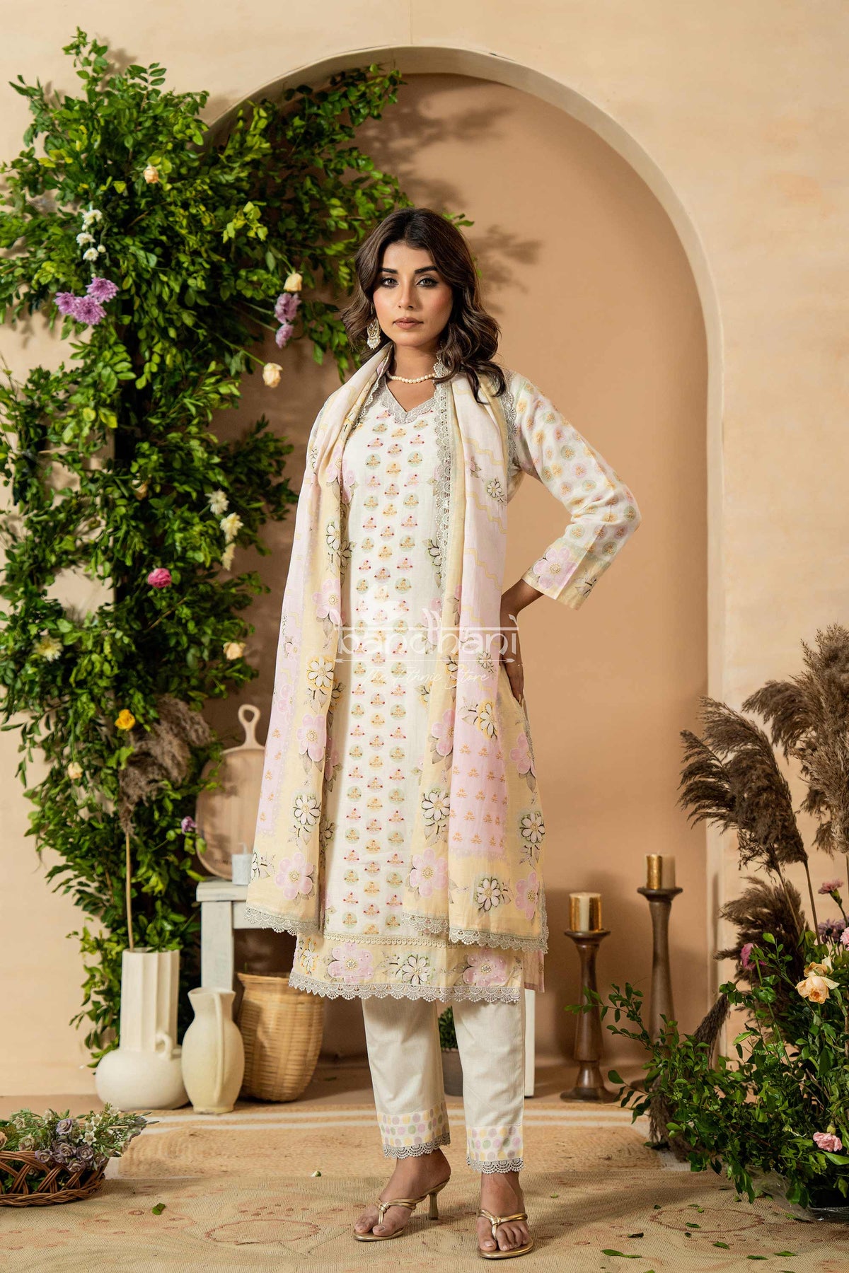 Woman in a floral embroidered outfit standing in a decorated indoor setting with plants and candles.