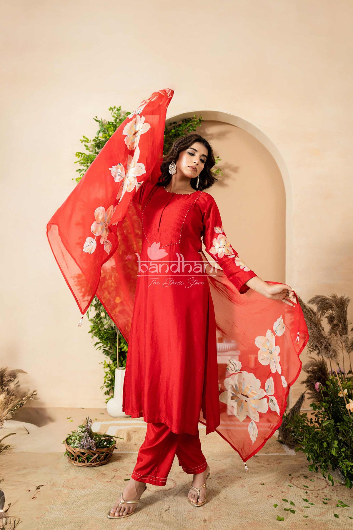 Woman in a red outfit with floral patterns holding a matching dupatta against a beige wall.