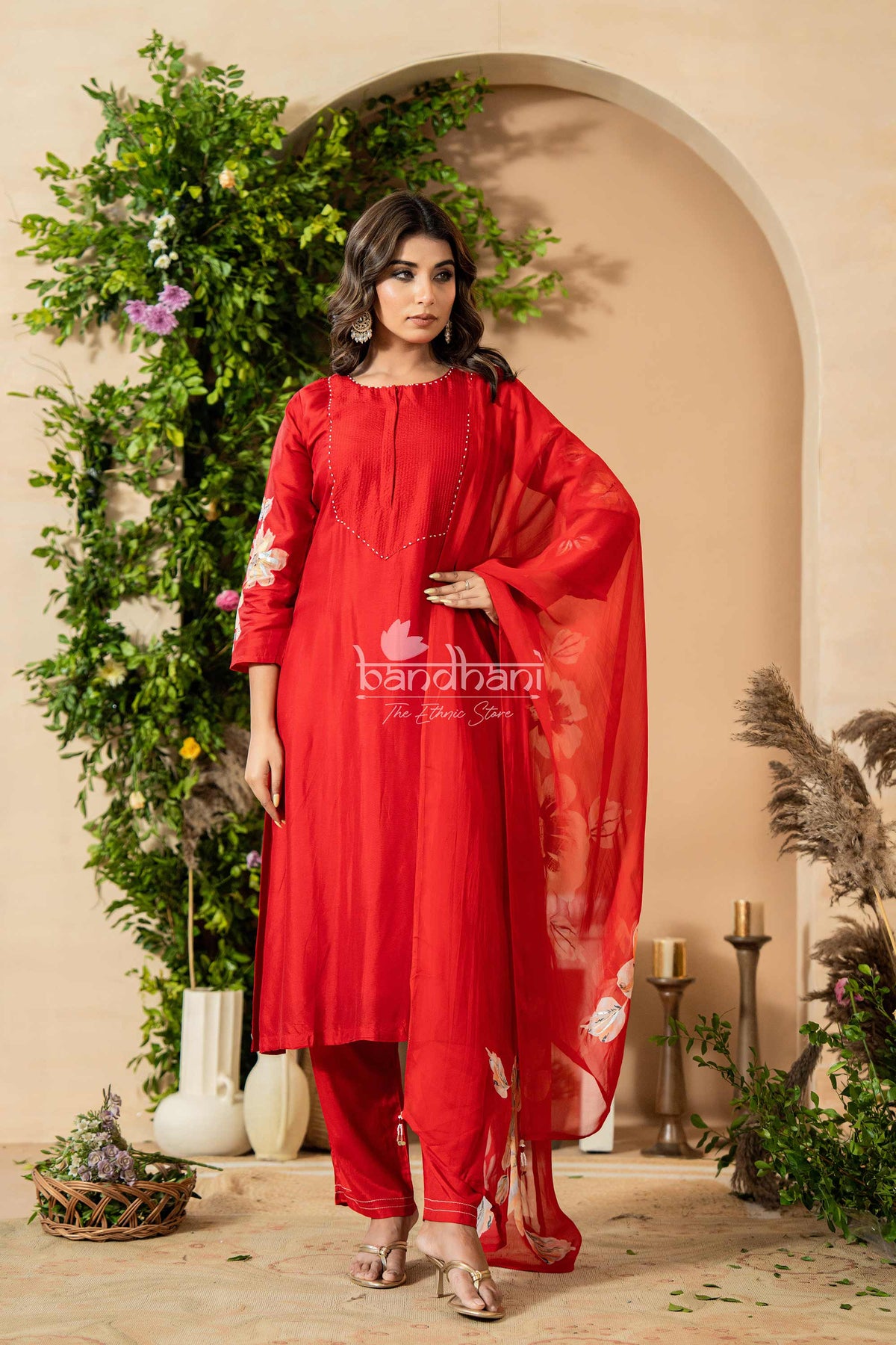 Woman wearing a red traditional outfit with 'bandhani' branding in a decorated indoor setting.