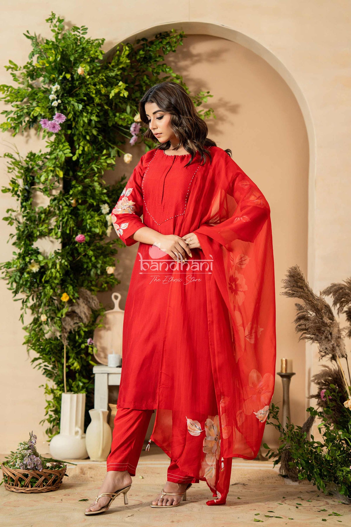 Woman in a red traditional outfit standing in a decorated indoor setting with plants and candles.