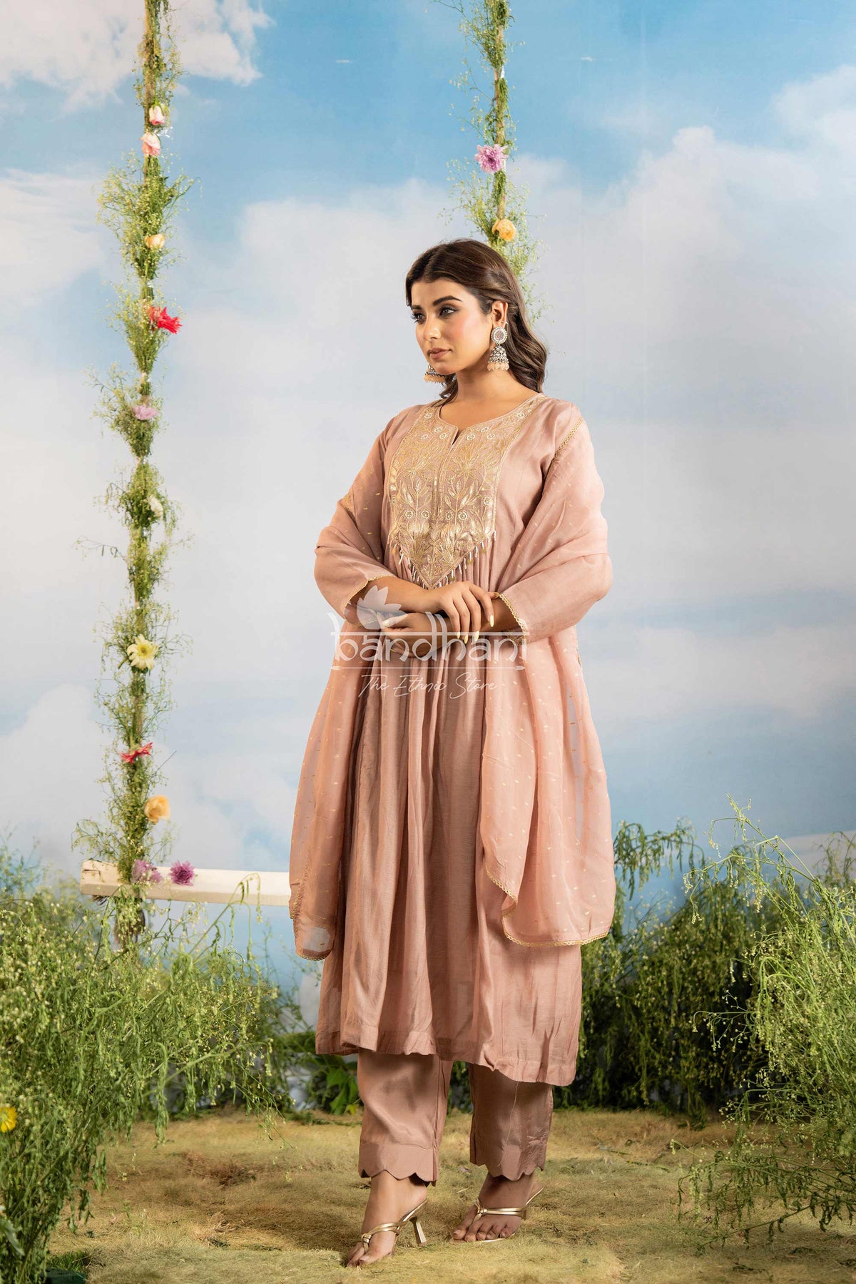 Woman in a pink traditional outfit standing outdoors with floral decorations and greenery.
