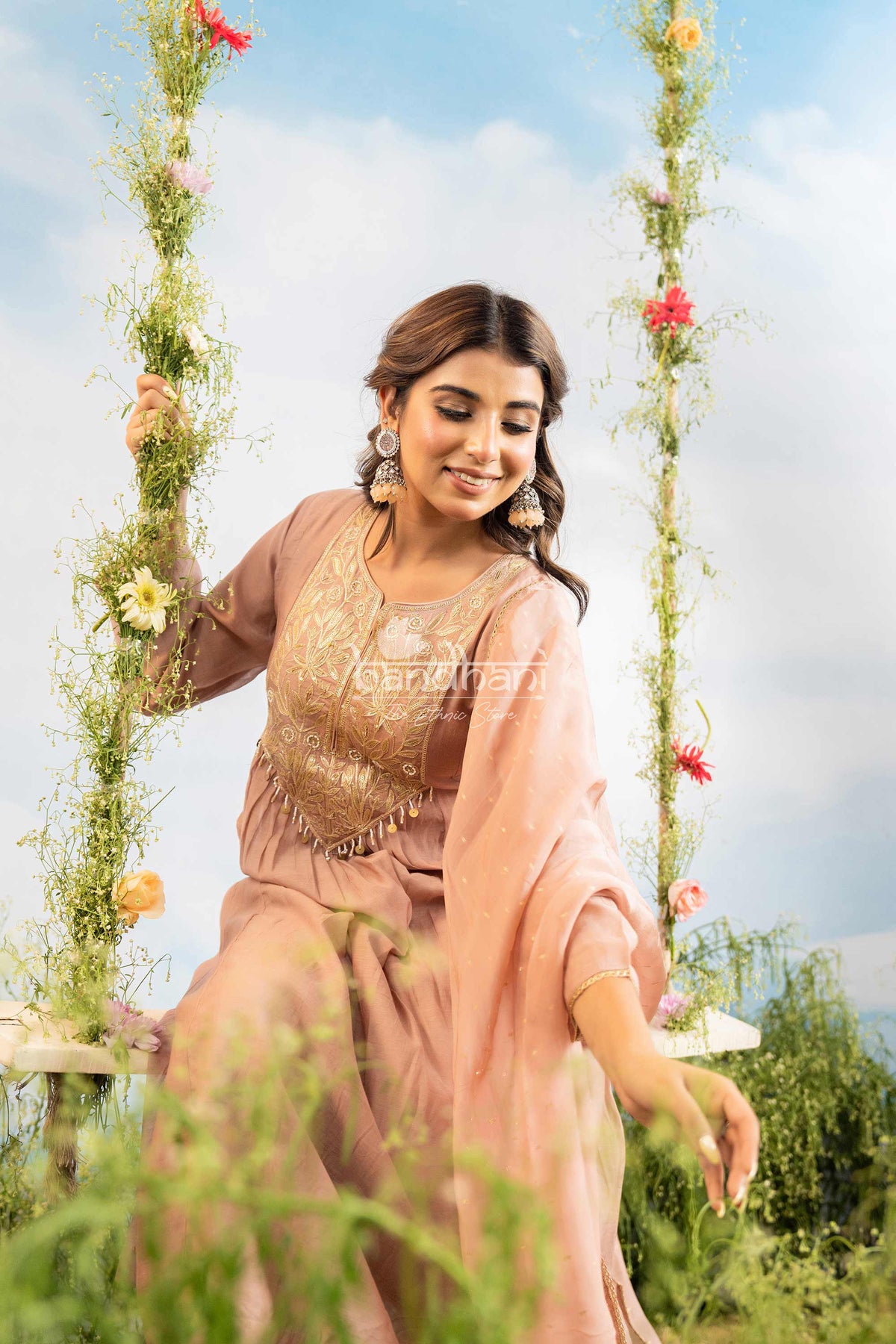 Woman in a pink and gold traditional outfit sitting on a floral swing with a scenic background.