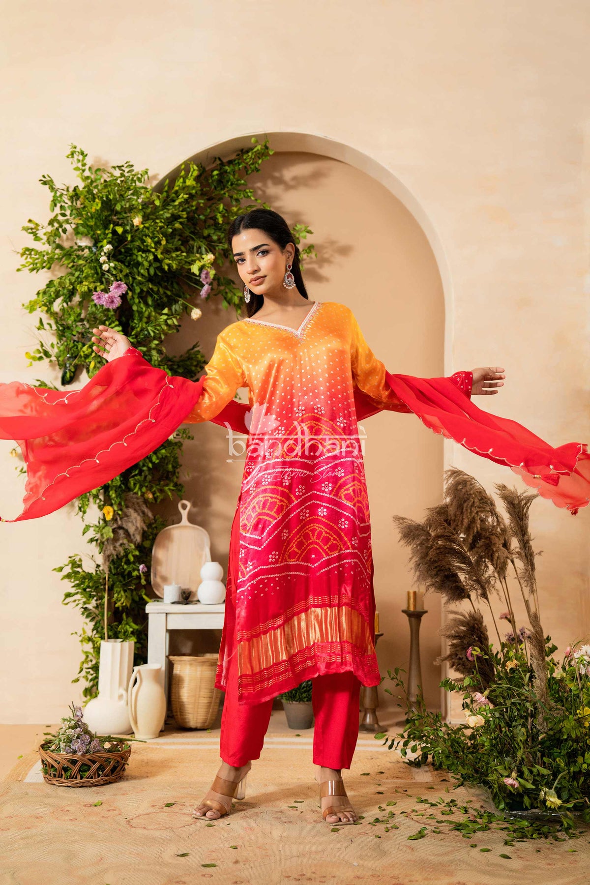 Woman in a colorful traditional outfit with a red and pink patterned dress, standing in a decorated room with plants and decor.