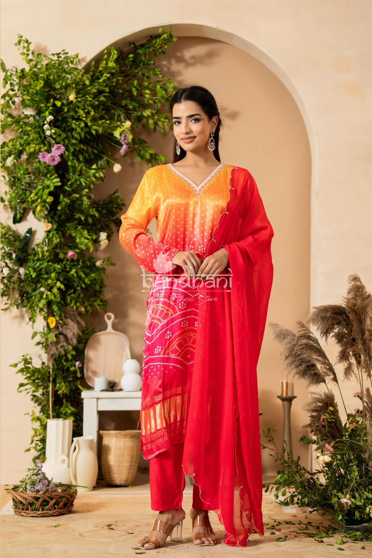 Woman in a red and orange traditional outfit standing in a decorated indoor setting with plants.