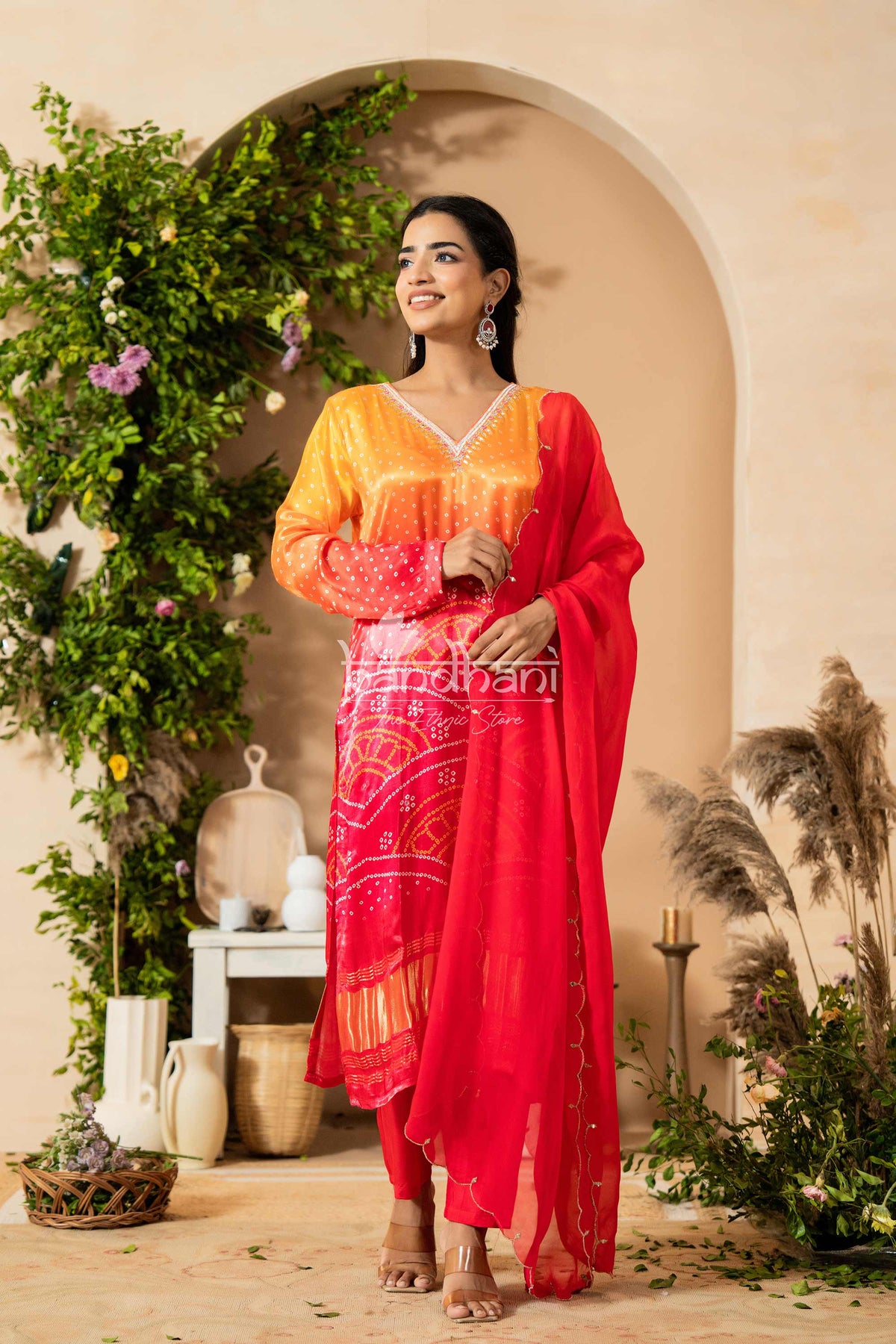 Woman in a red and orange traditional outfit standing in a decorated indoor setting with plants and archway.