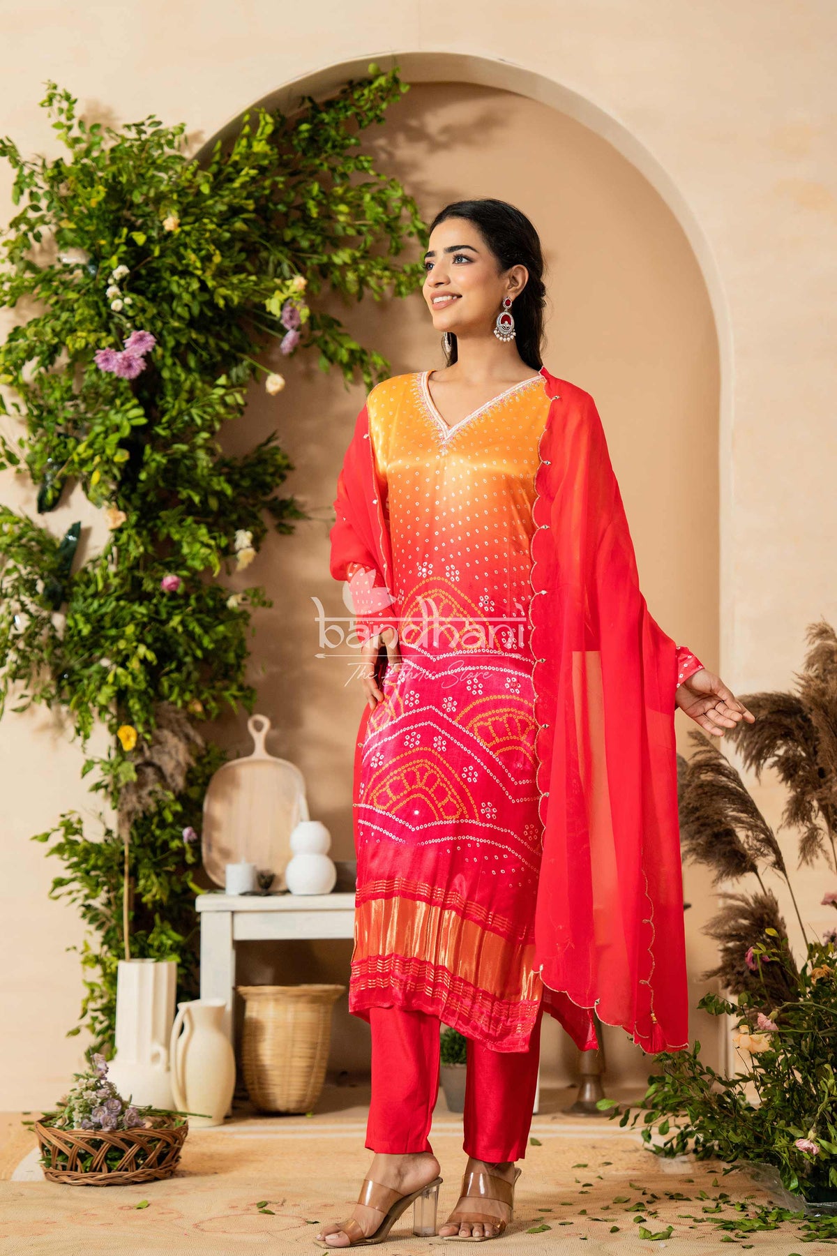 Woman in a red and orange traditional outfit standing in a decorated indoor setting with plants and decor.