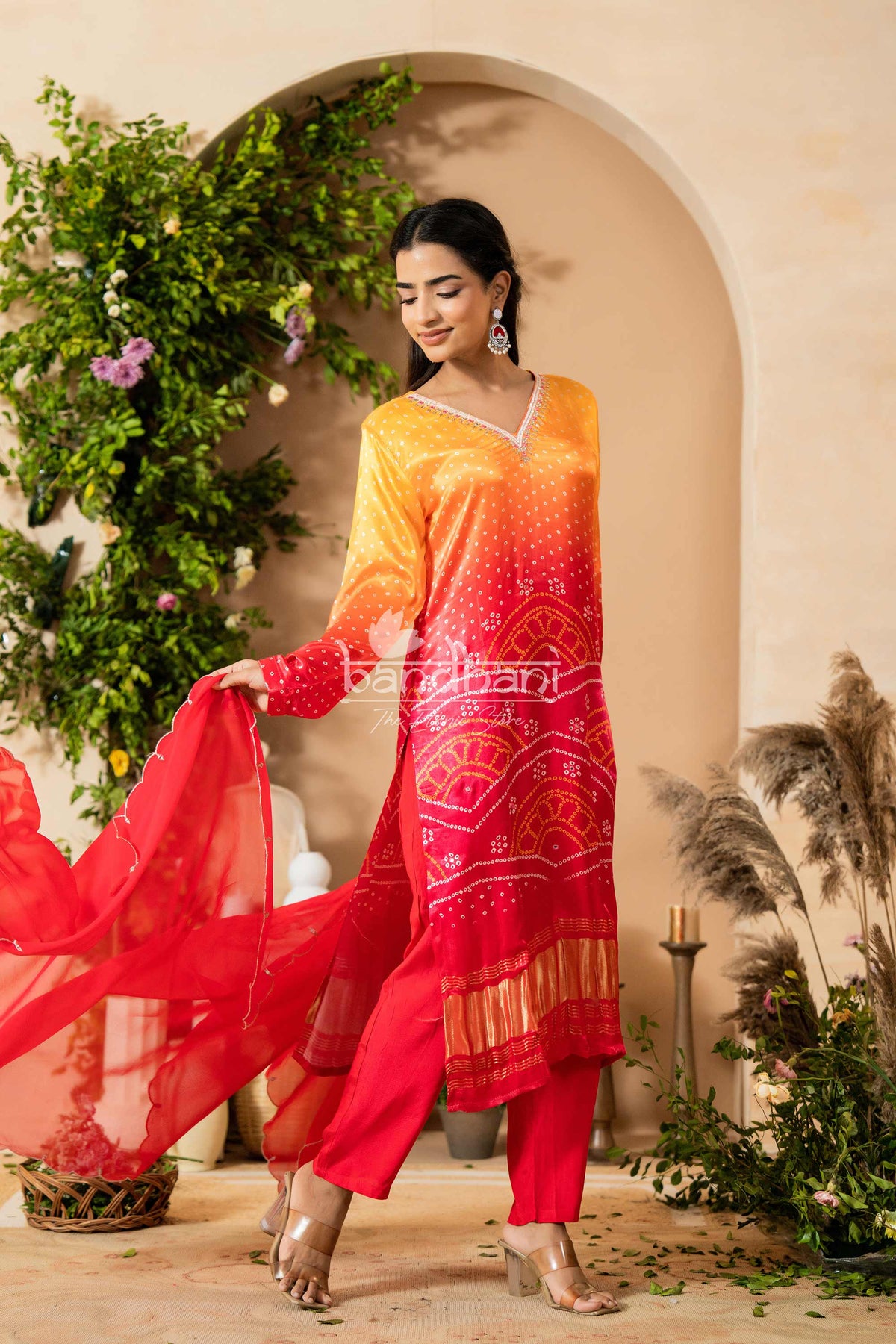 Woman in a red and orange traditional outfit with 'Bharat Bani' branding, standing in a decorative setting.