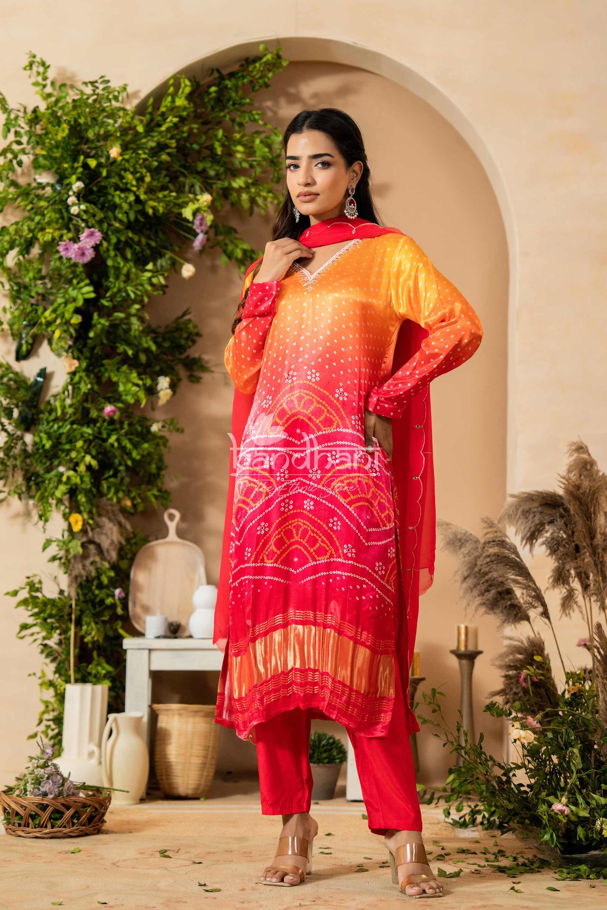 Woman in a red and orange traditional outfit standing in a decorative indoor setting with plants.