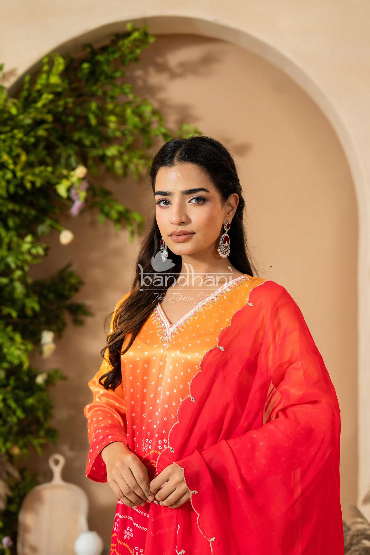 Woman in a red and orange traditional outfit standing in front of an archway with greenery.