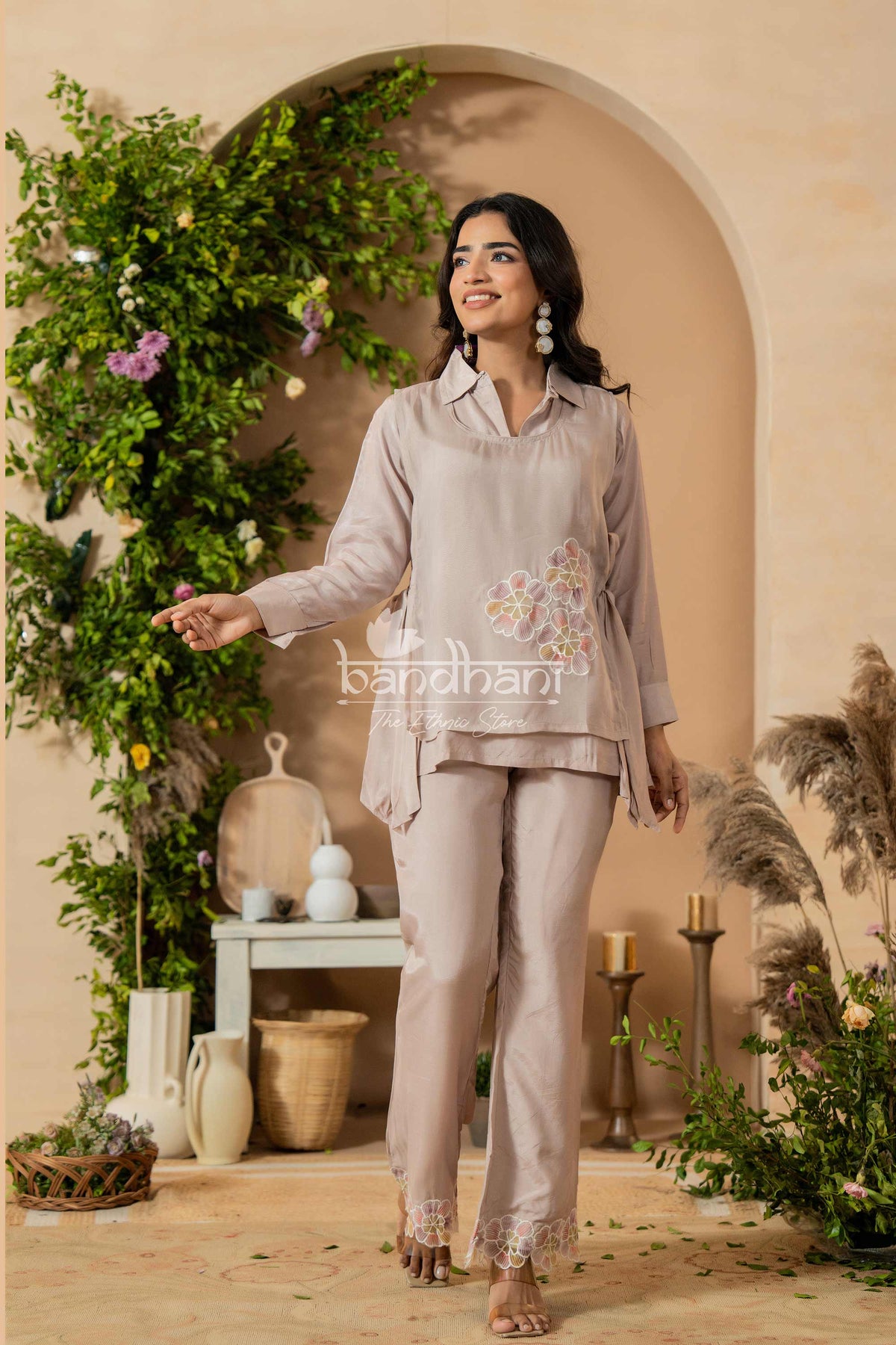 Woman in a beige outfit with floral embroidery standing in a decorated room with plants and candles.