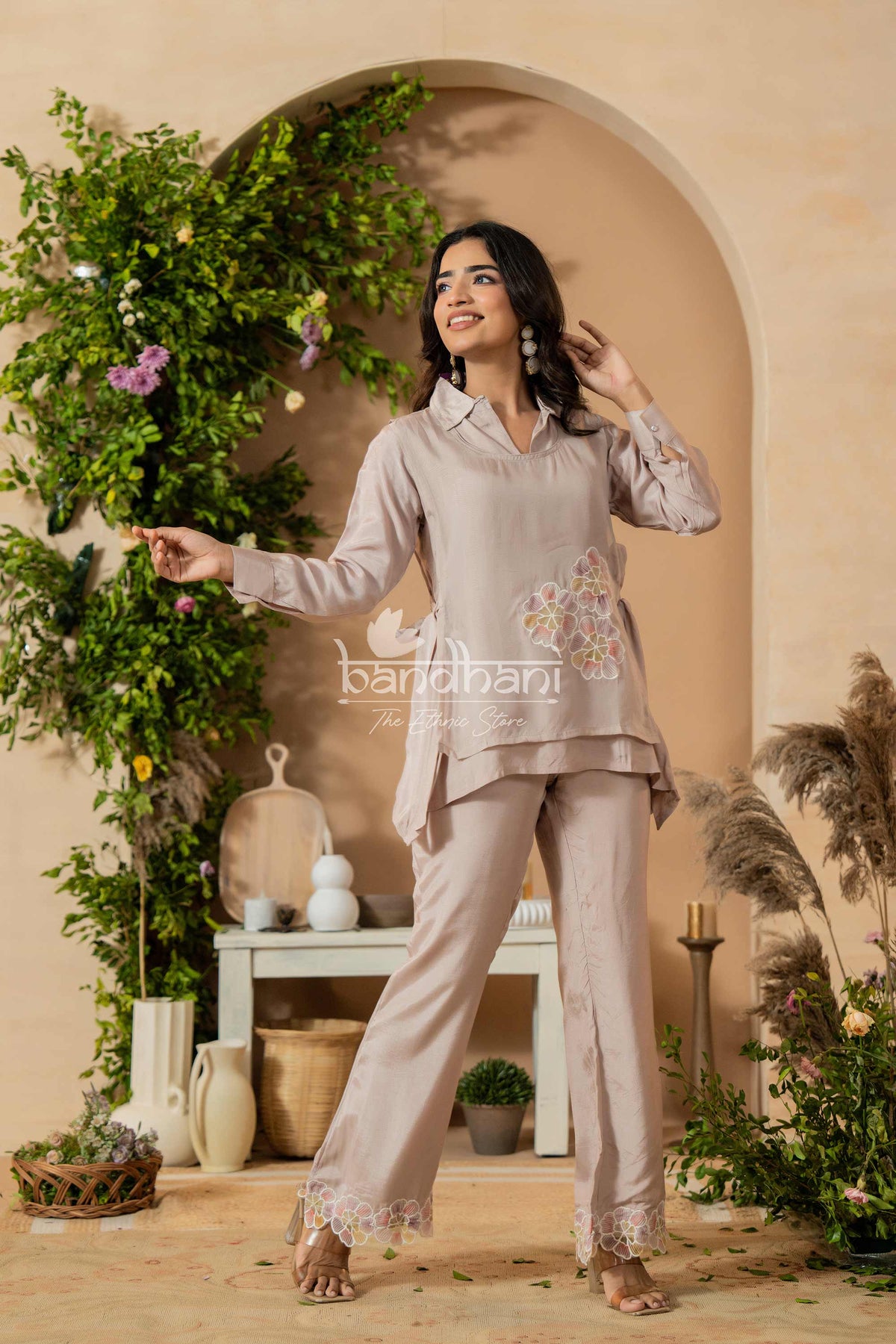Woman in a beige outfit standing in a decorated indoor setting with plants and archway.