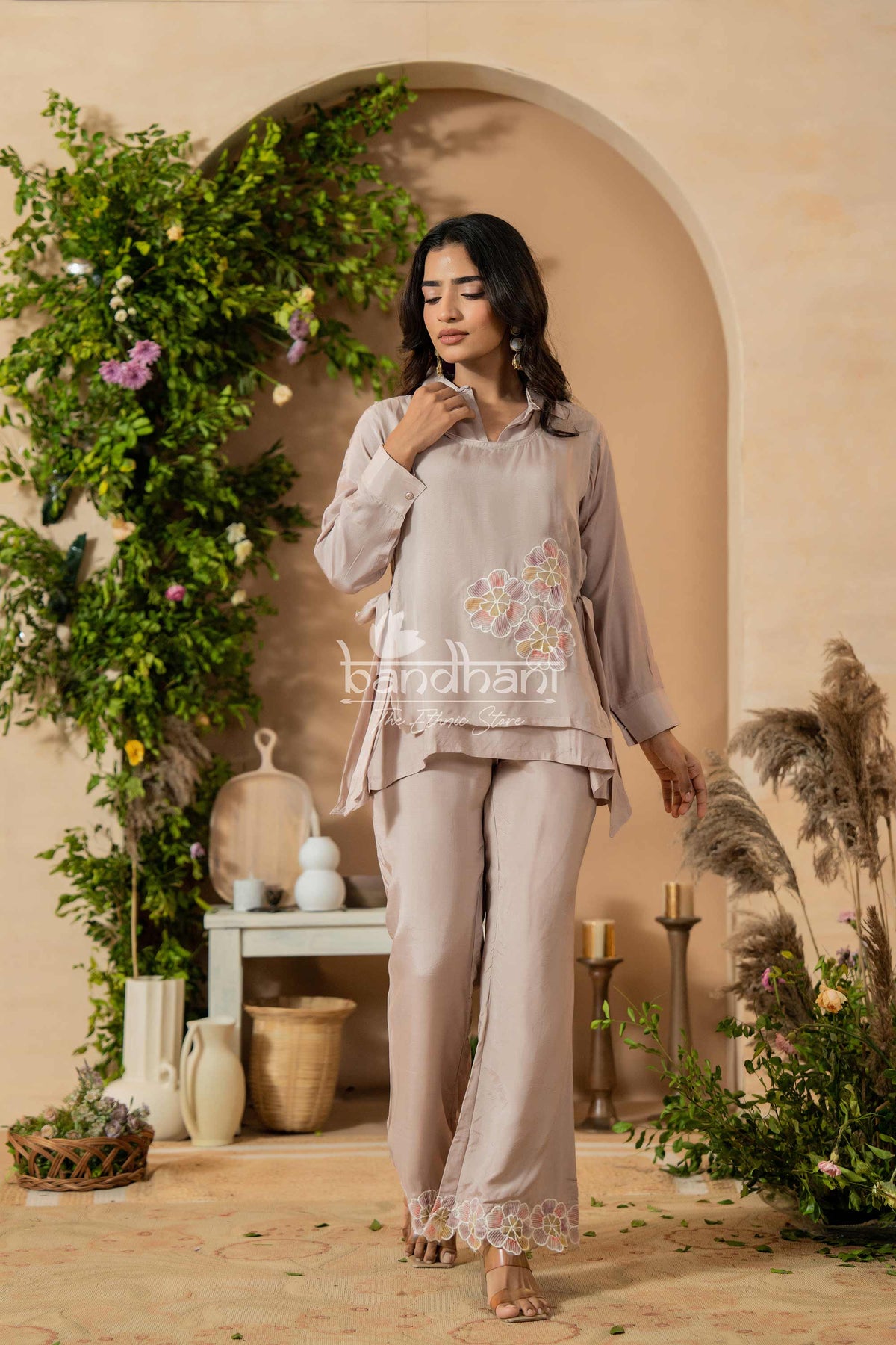 Woman in a light gray traditional outfit with floral embroidery standing in a decorated indoor setting.