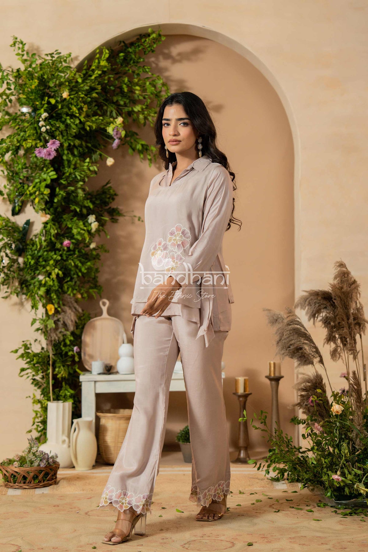 Woman in a light gray outfit standing in a decorated indoor setting with plants and archway.
