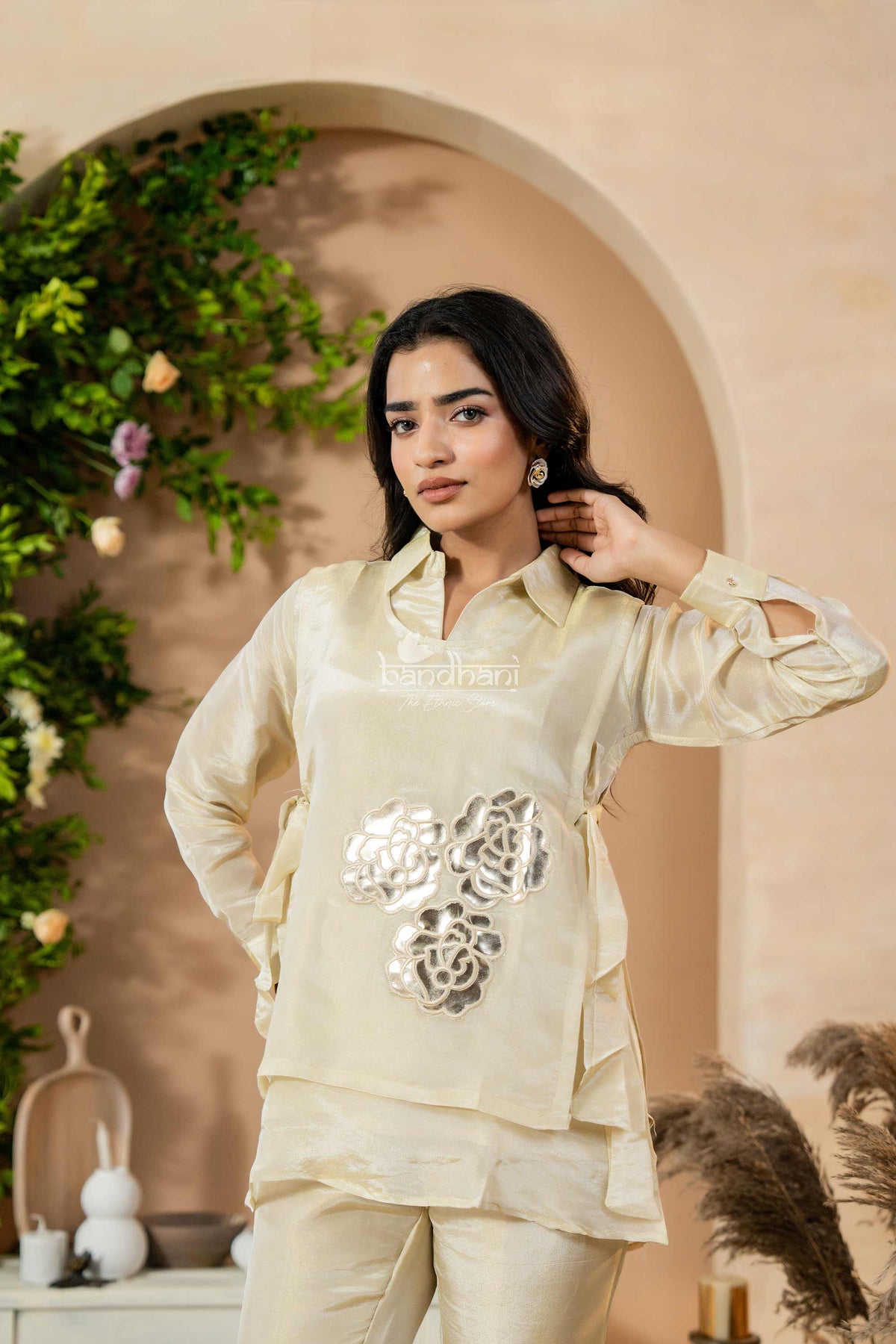 Woman in a cream traditional outfit with floral embroidery standing in front of an archway with greenery.