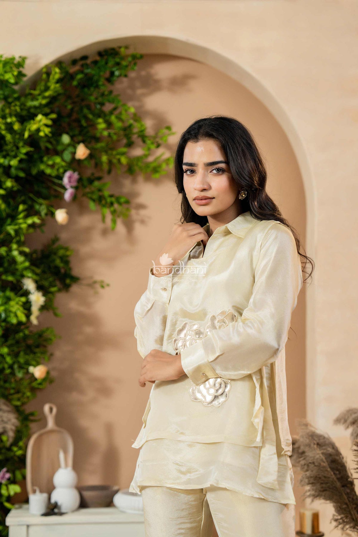 Woman in a cream-colored traditional outfit standing in front of an archway with greenery.
