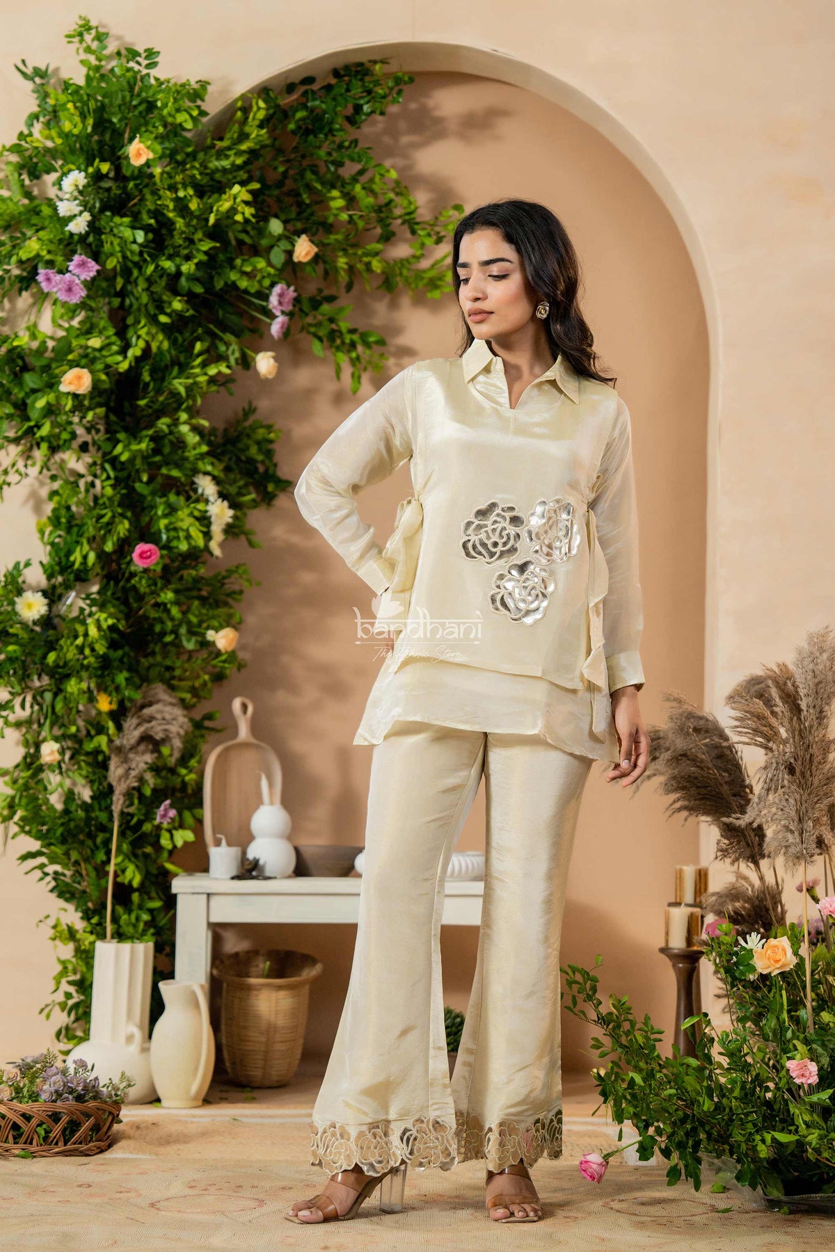 Woman in a cream outfit standing in a decorated room with plants and flowers.