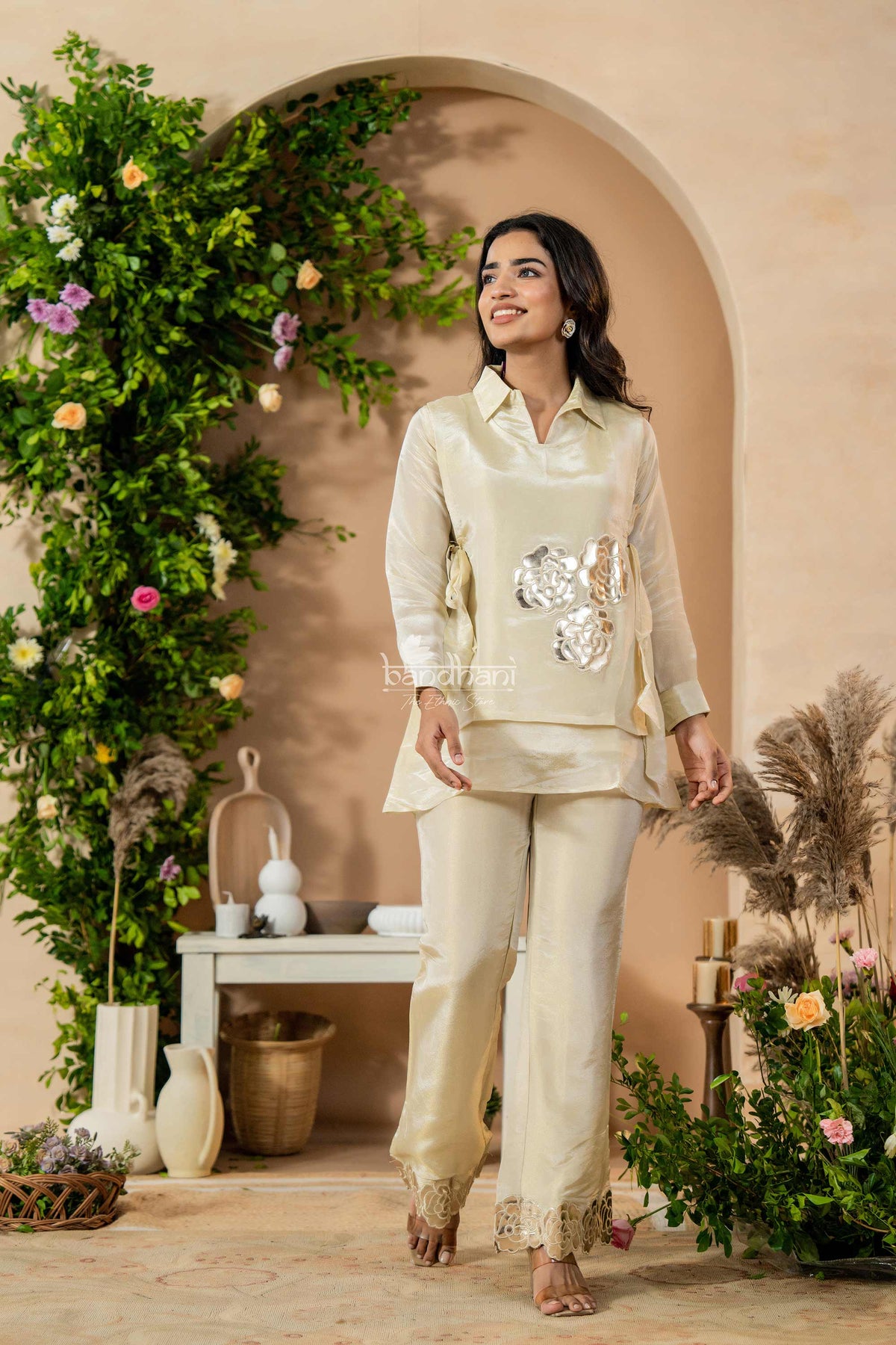 Woman in a cream traditional outfit standing in a decorated indoor setting with plants and flowers.