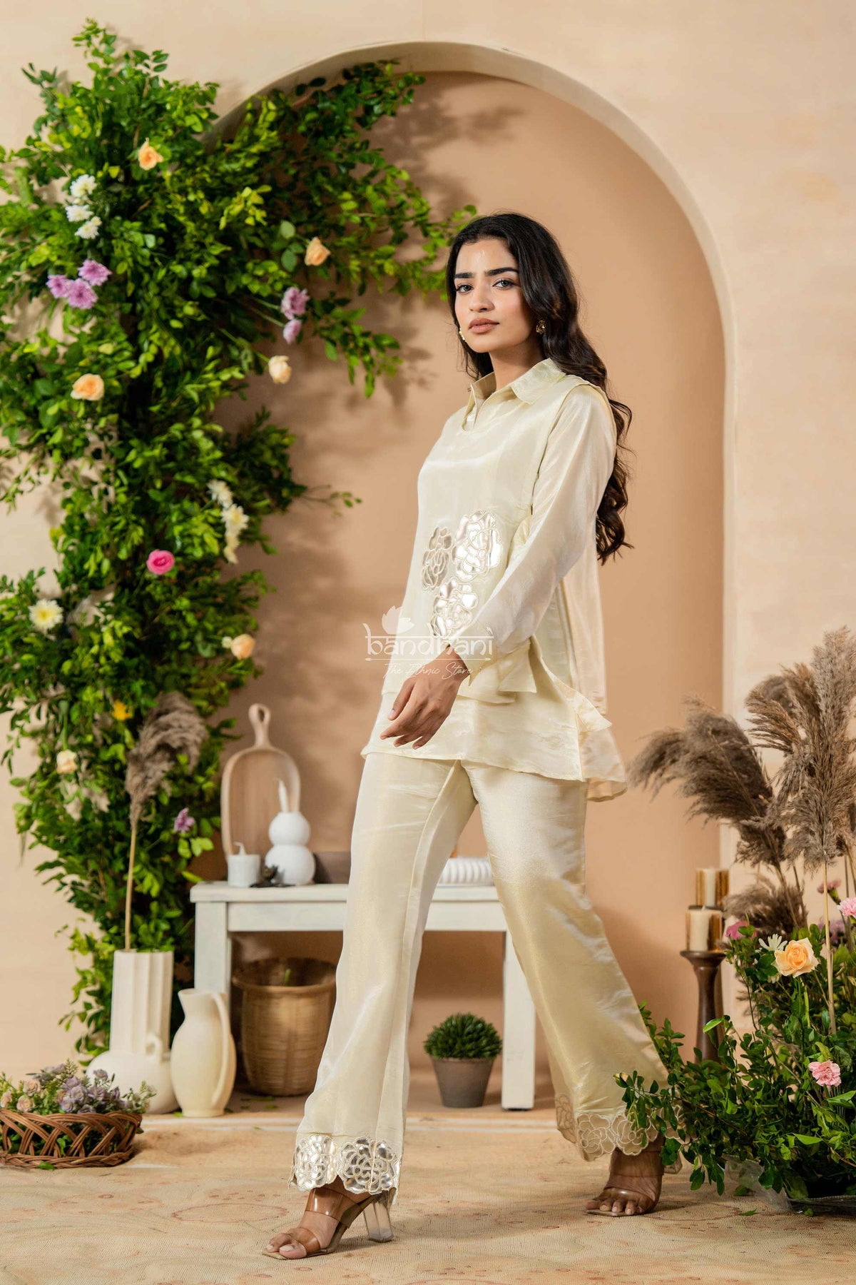 Woman in a white traditional outfit standing in a decorated indoor setting with plants and flowers.