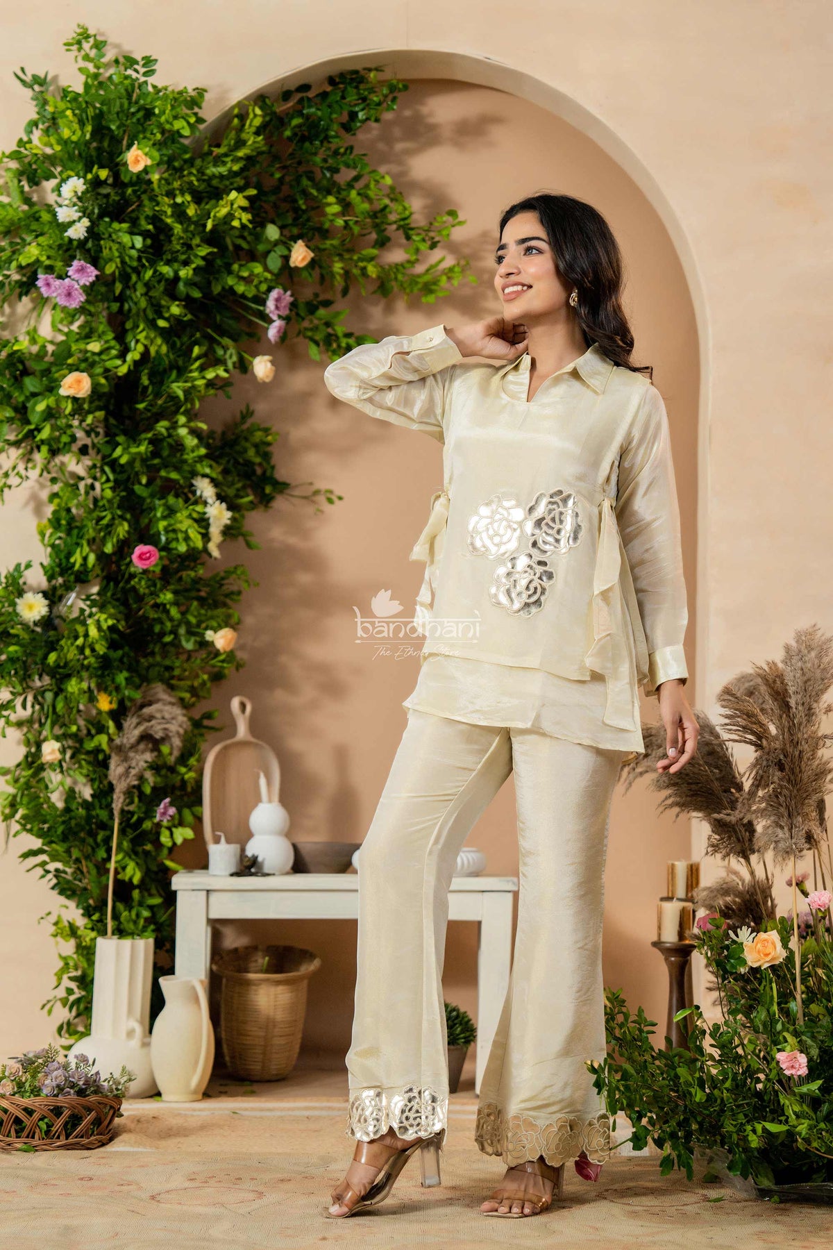 Woman in a beige traditional outfit standing in a decorated indoor setting with flowers and decor.