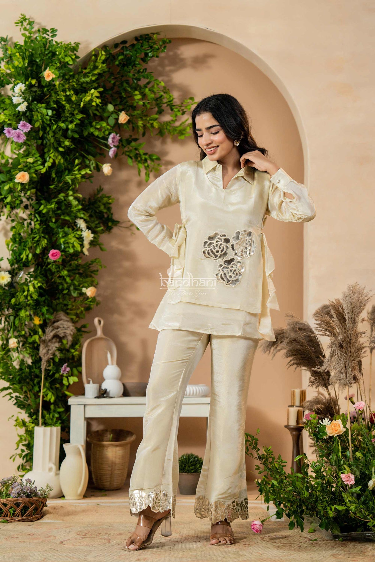 Woman in a cream traditional outfit standing in a decorated indoor setting with plants and flowers.