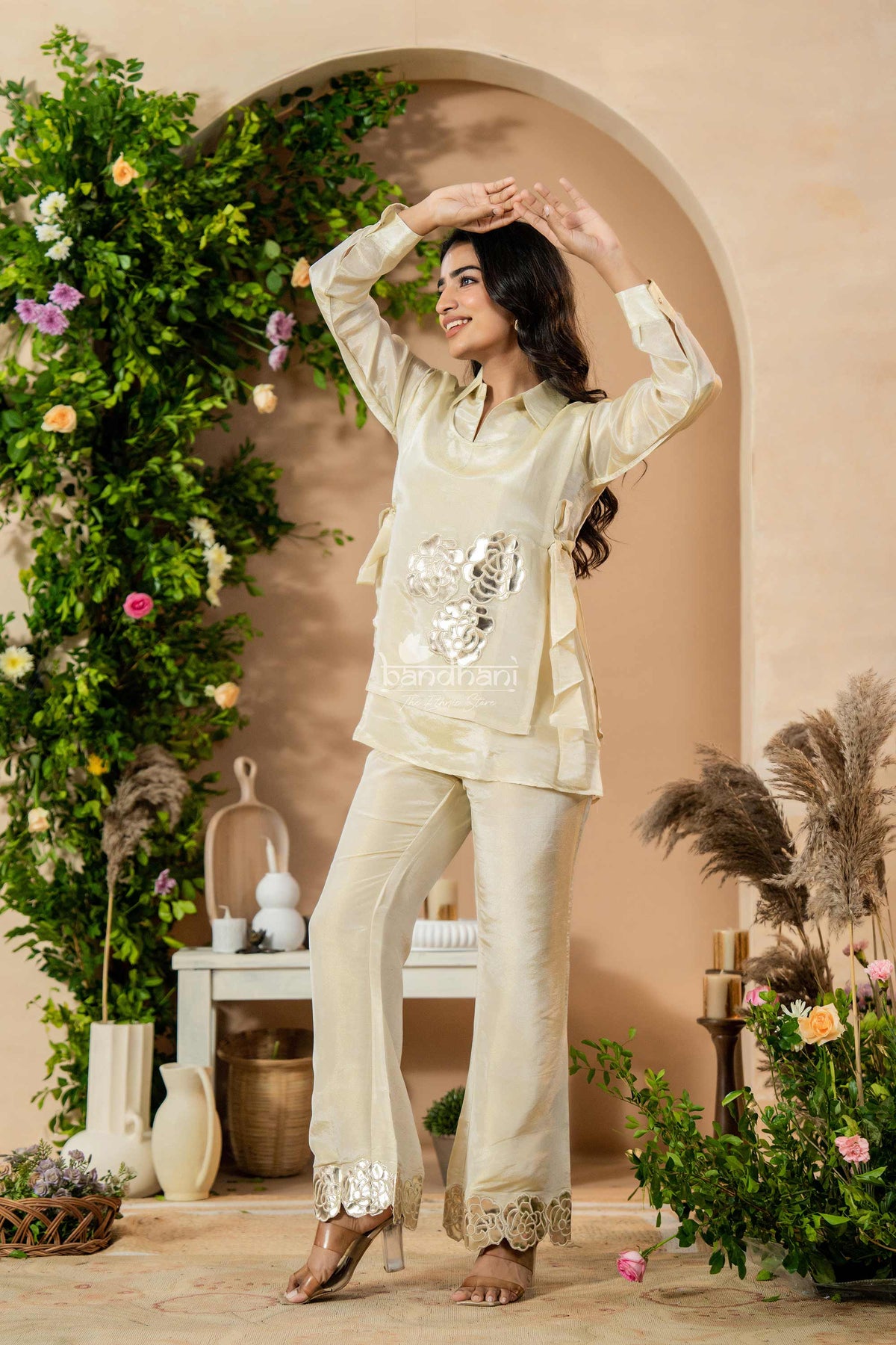 Woman in a cream traditional outfit standing in a decorated room with plants and decor.
