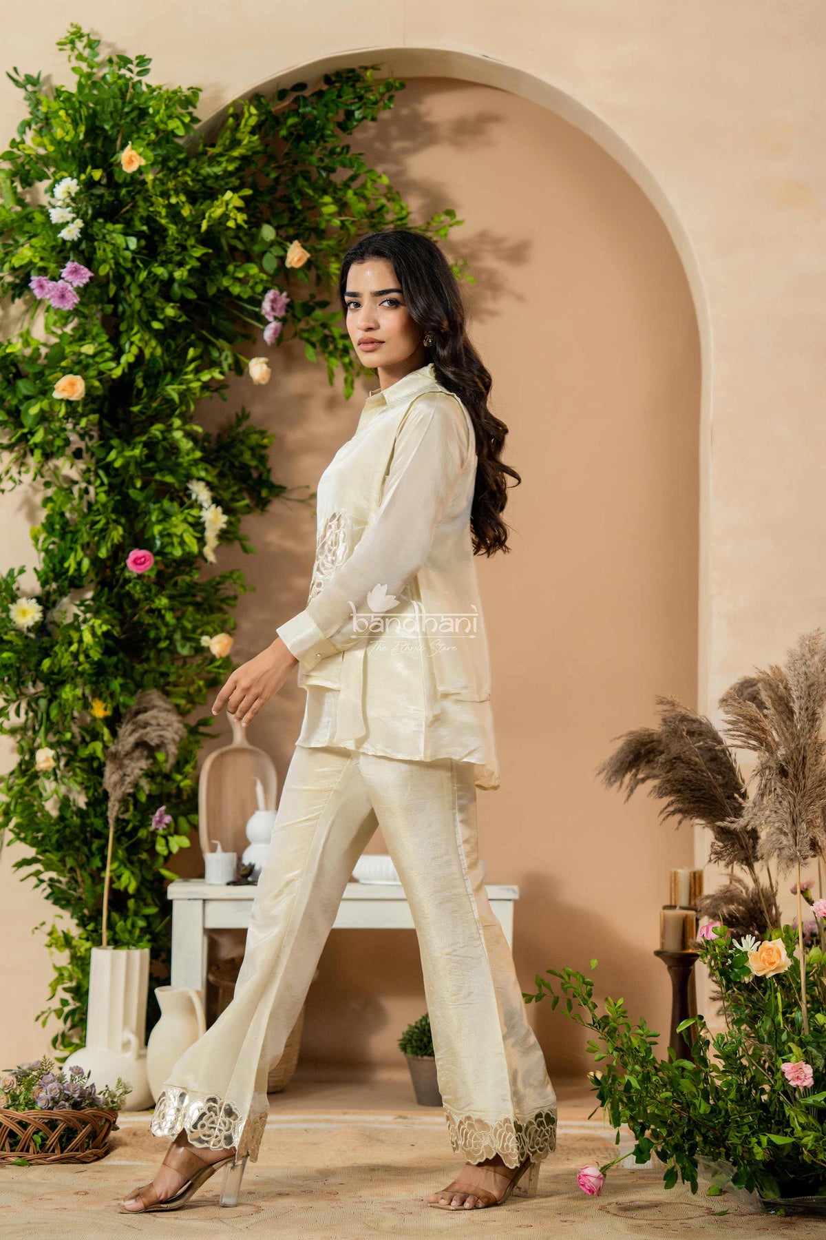 Woman in a white outfit standing in front of a decorated archway with flowers and plants.