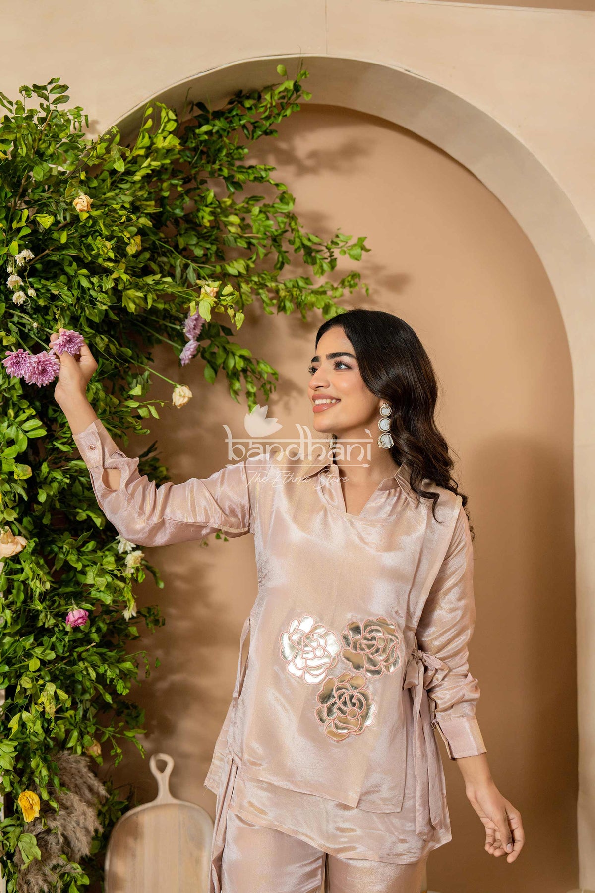 Woman in a light pink traditional outfit standing next to a decorative wreath with flowers.