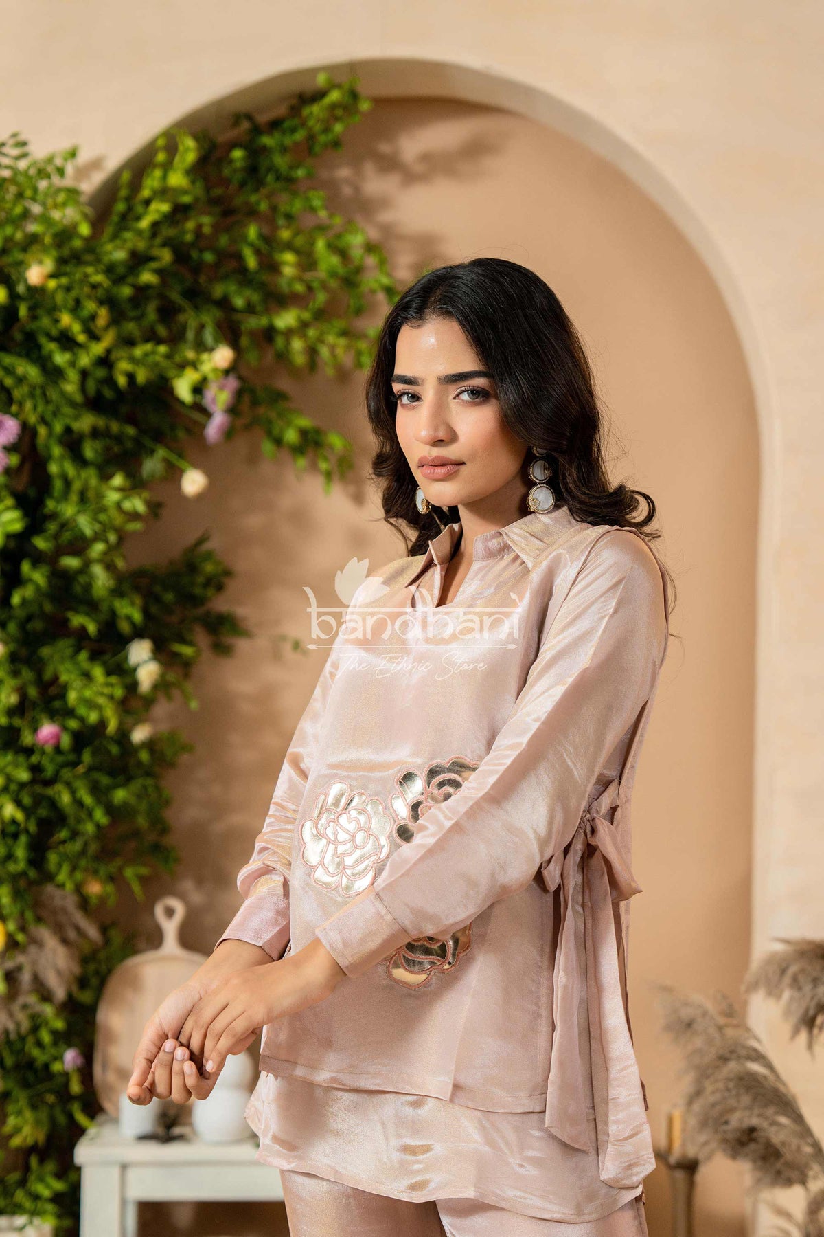 Woman in a light pink outfit standing in a decorative indoor setting with plants and archway.