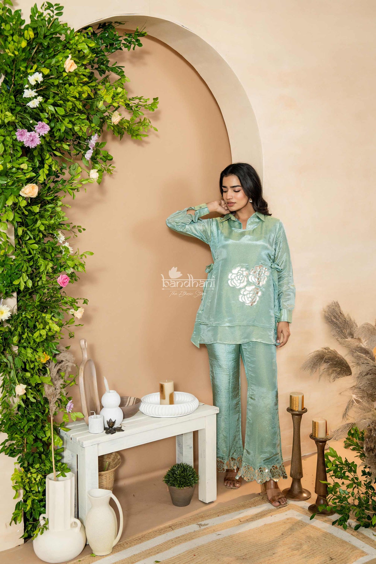 Woman in a light green traditional outfit standing in a decorated room with plants and candles.