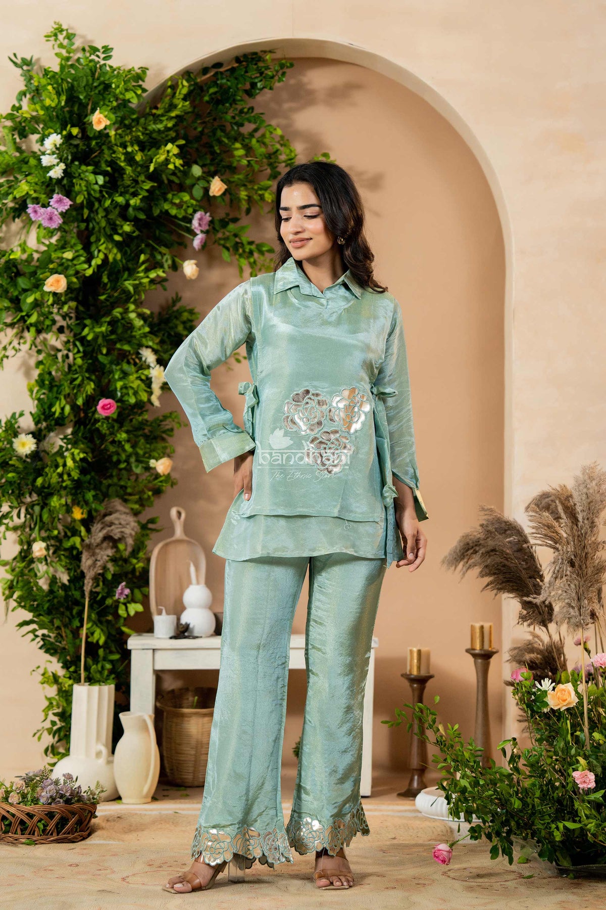 Woman in a light blue traditional outfit standing in a decorated indoor setting with plants and flowers.
