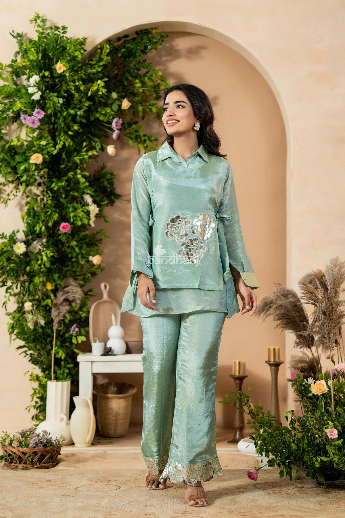 Woman in a light green traditional outfit standing in a decorated indoor setting with flowers and plants.
