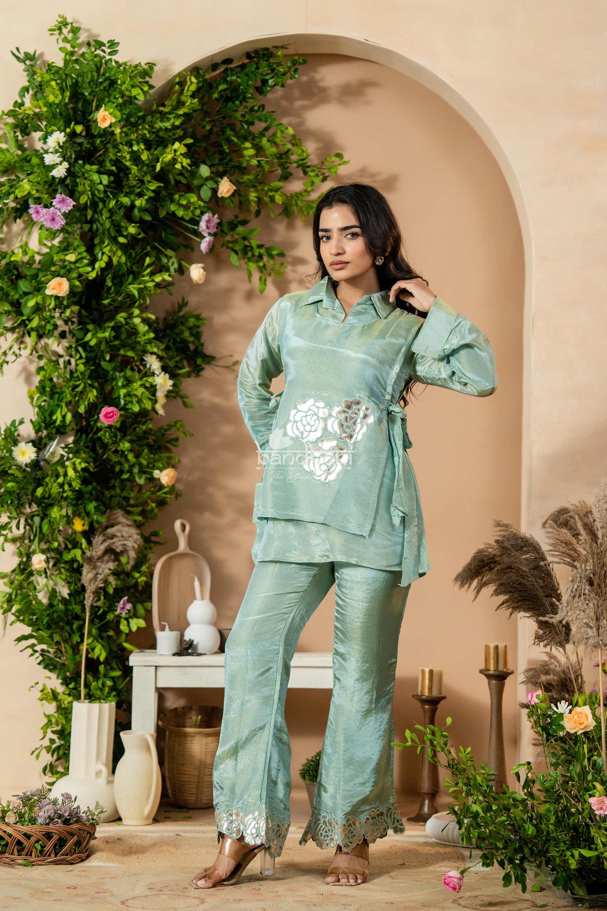 Woman in a light green traditional outfit standing in a decorated indoor setting with plants and flowers.