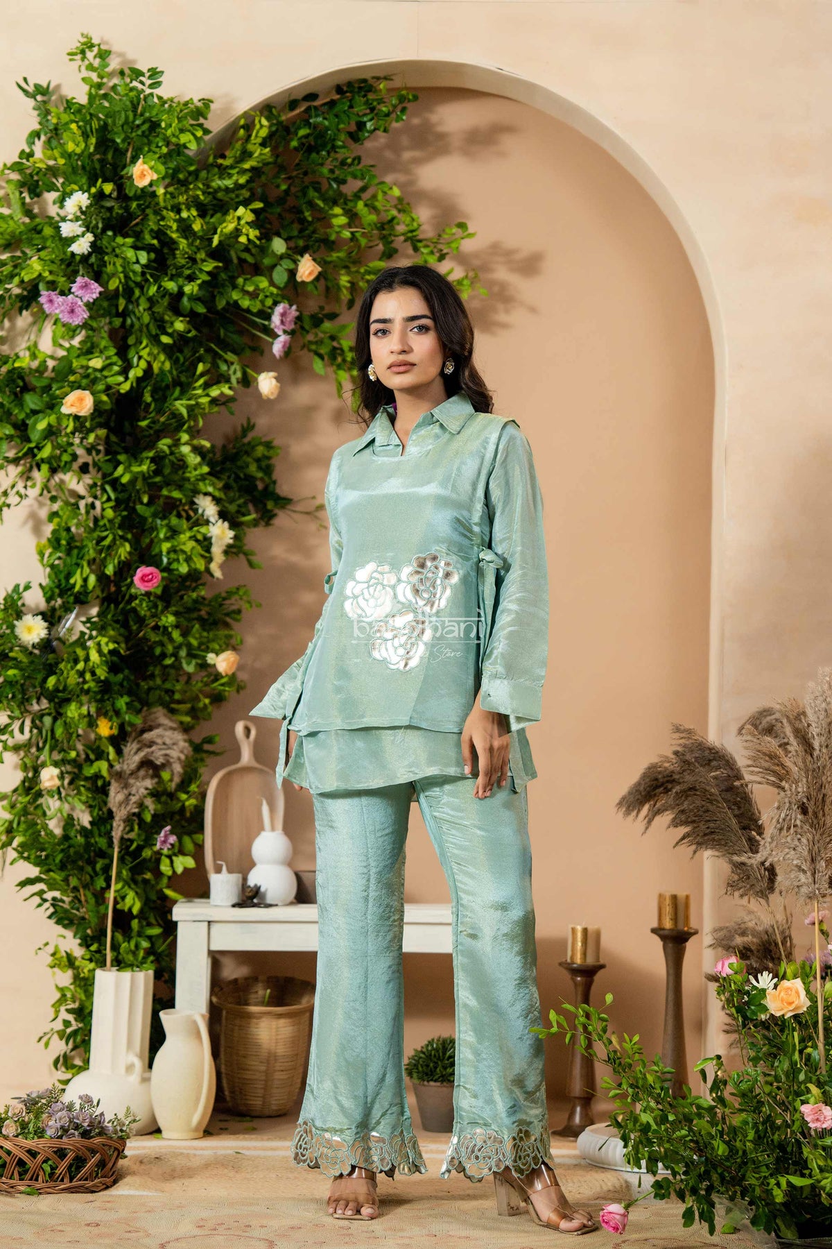 Woman in a light blue traditional outfit standing in a decorated indoor setting with plants and flowers.