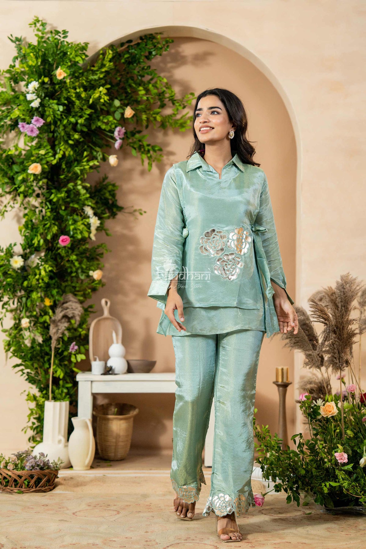 Woman in a light green traditional outfit standing in a decorated indoor setting with flowers and plants.