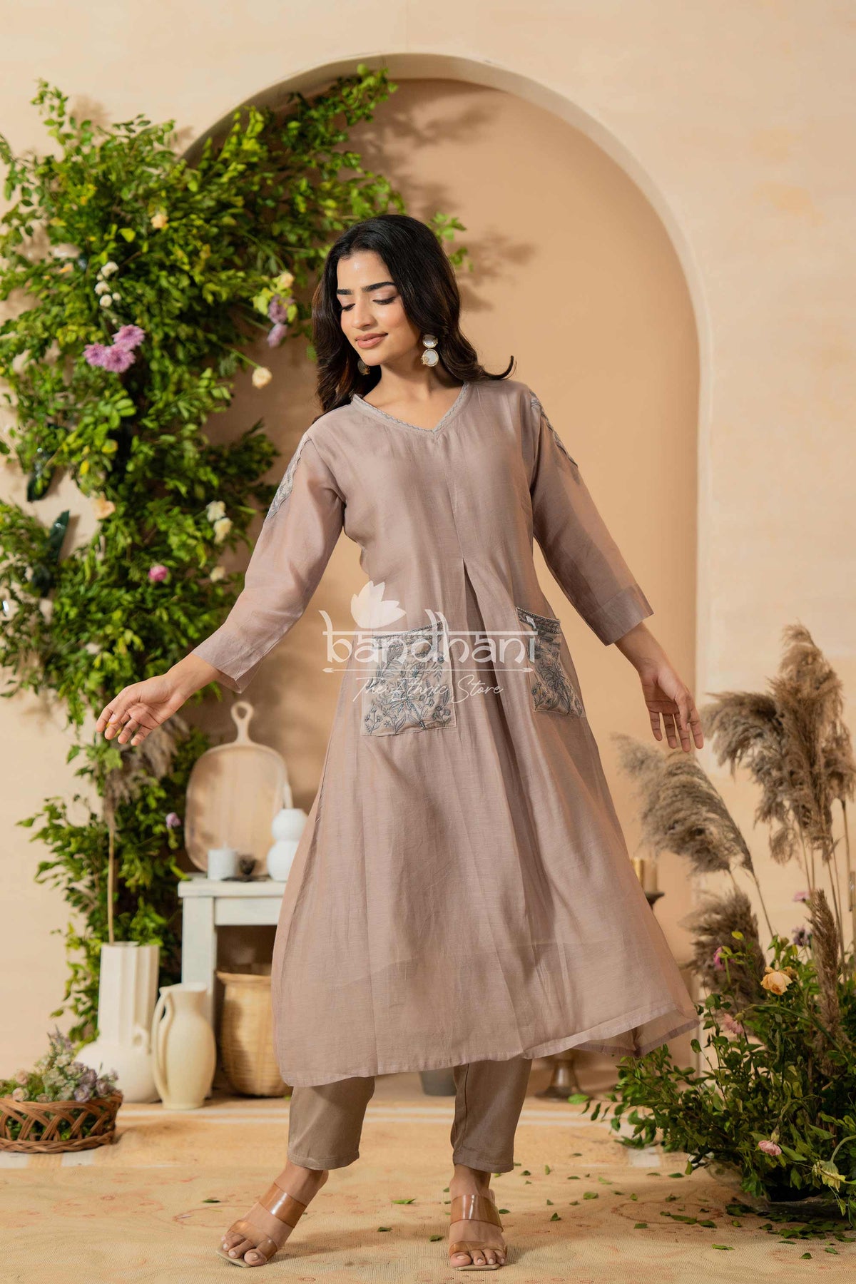 Woman in a light brown dress with floral details standing in a decorated indoor setting.