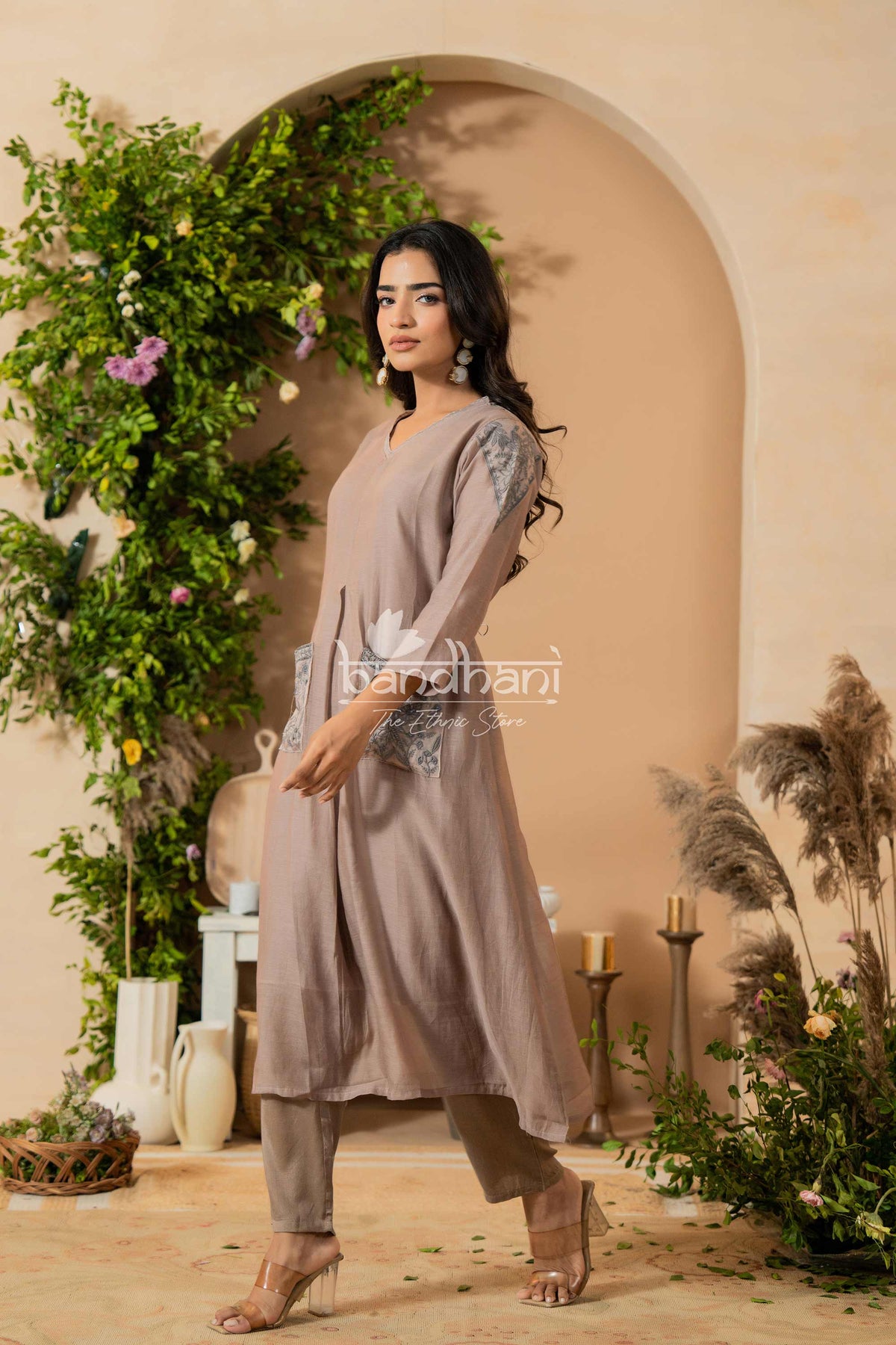Woman in a long, light-colored dress standing in a decorated indoor setting with plants and archway.