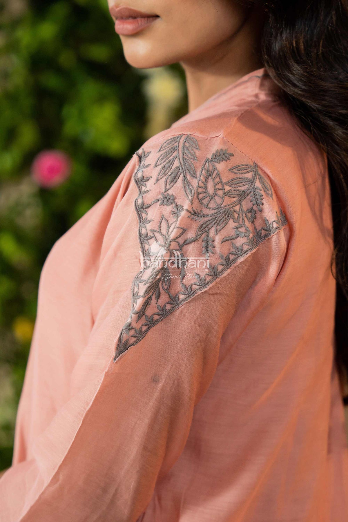 Person wearing a peach-colored garment with floral lace detailing on the shoulder, against a blurred green background.