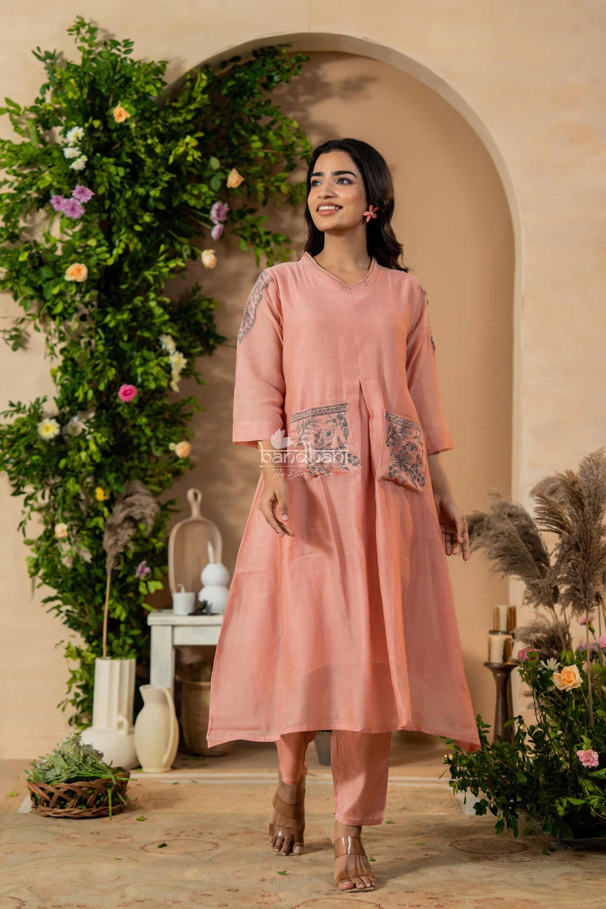 Woman in a pink traditional outfit standing in a decorated indoor setting with plants and flowers.