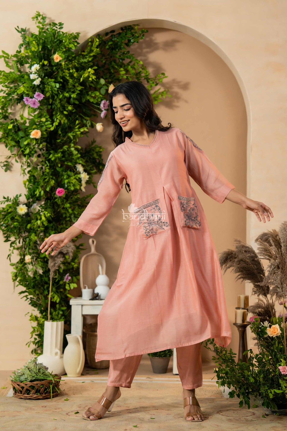 Woman in a pink traditional outfit standing in a decorated indoor setting with plants and flowers.