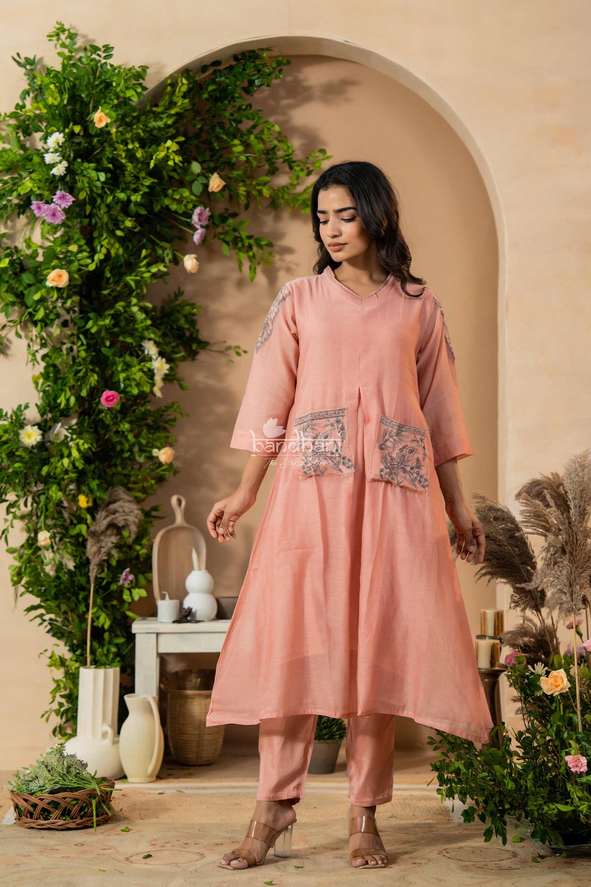Woman in a pink traditional outfit standing in a decorated indoor setting with greenery and flowers.