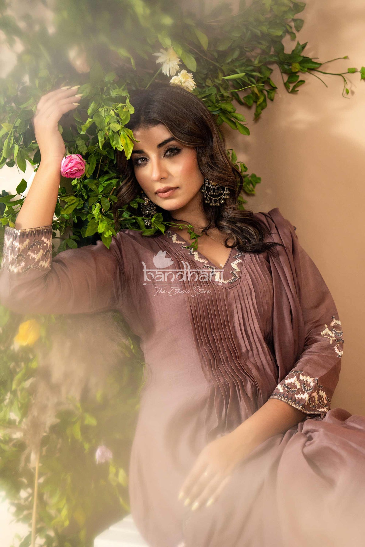 Woman in a brown and white outfit with 'bandhani' branding, standing against a green leafy background.